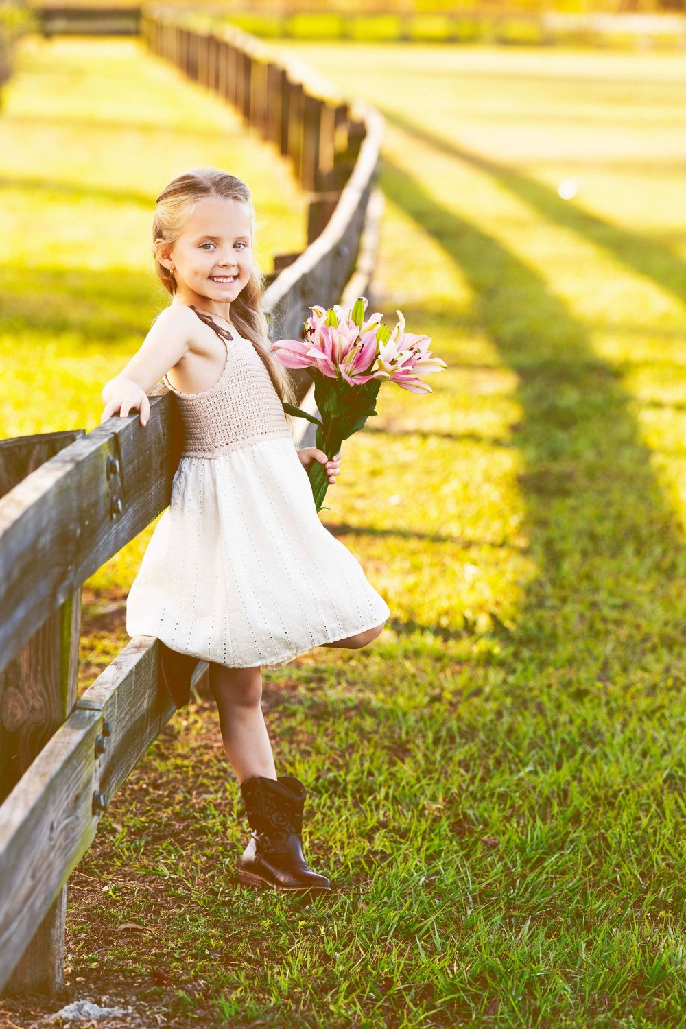 Child in white dress holding pink lilies by rustic fence in sunny field.