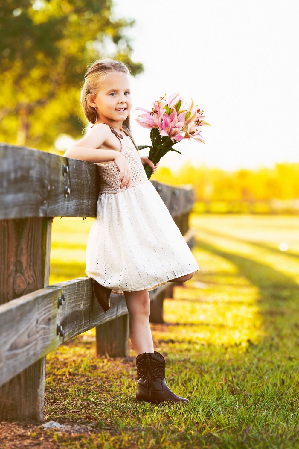 Person in tan and white dress holding pink lilies while leaning on wooden fence at sunset.