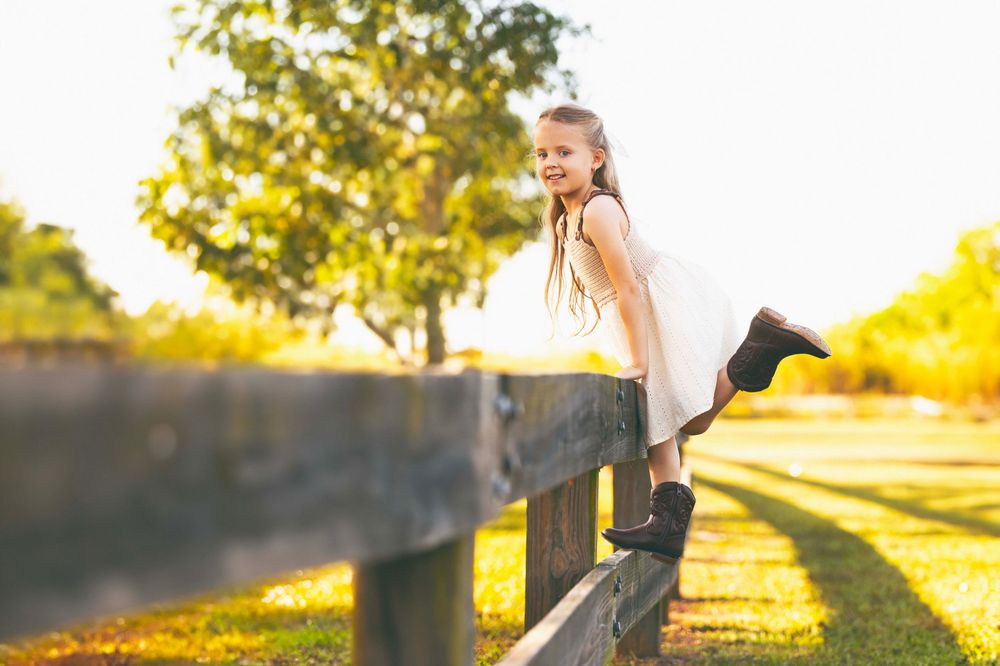 Young figure in white dress balancing playfully on wooden fence during golden sunset in park.