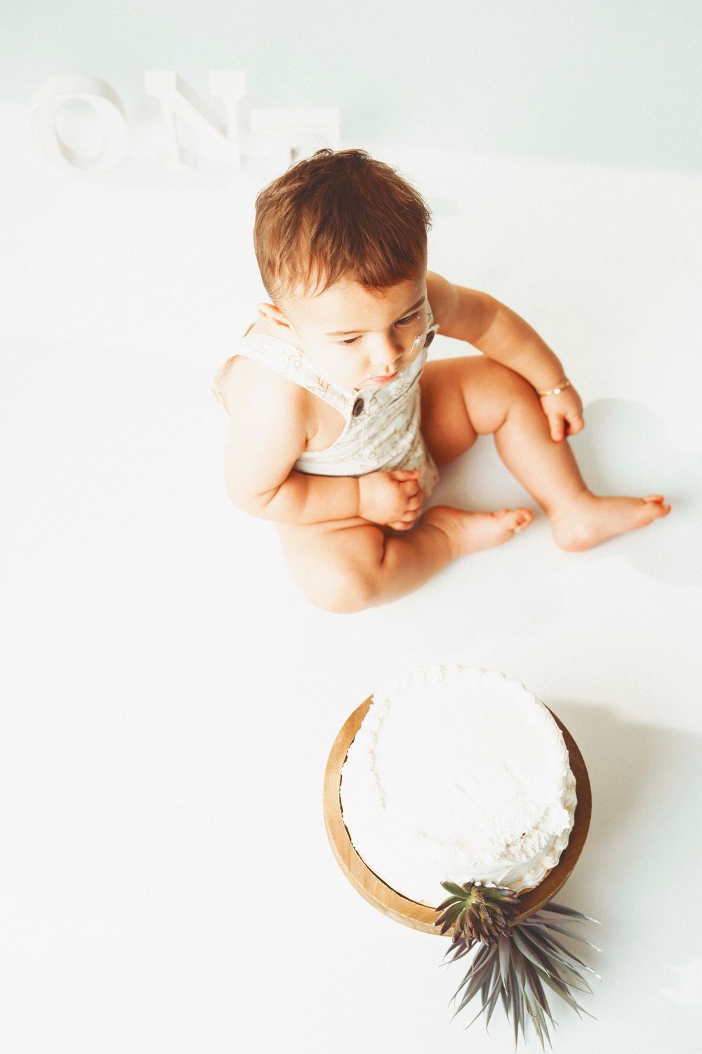 Baby in white onesie sitting next to a white decorated cake with greenery on a light background.
