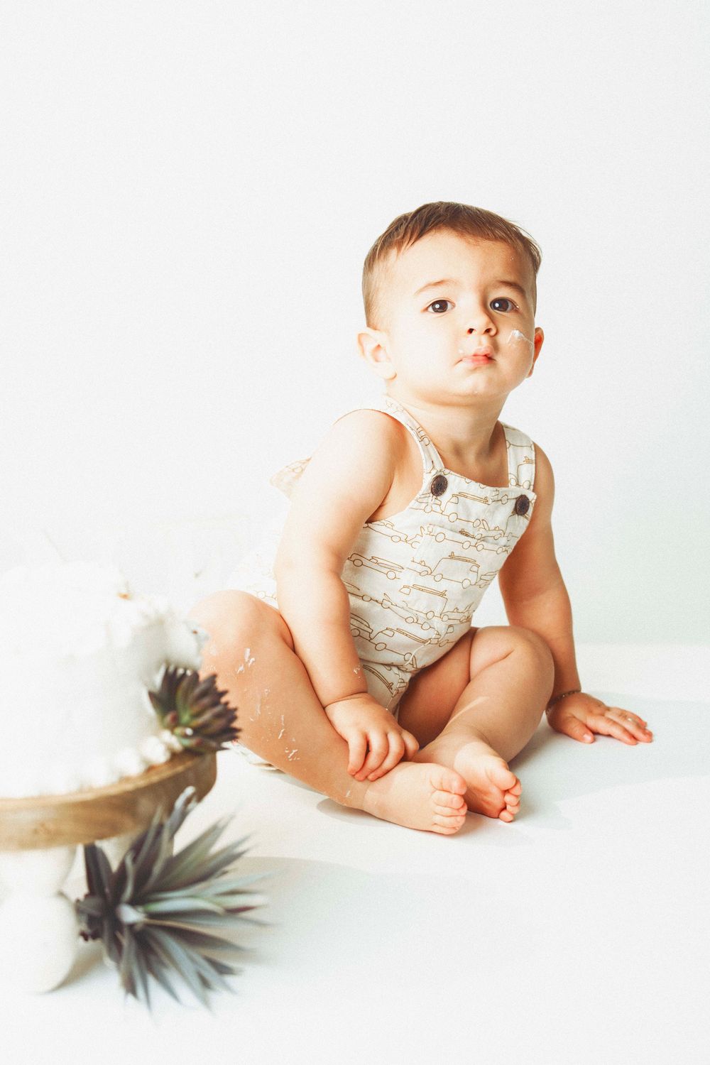 Baby in white romper sitting on light background next to decorative cake.