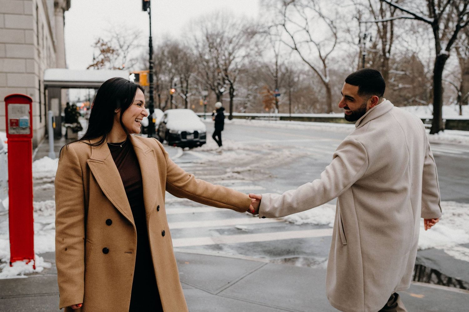 Playful couple holding hands on snowy city sidewalk in winter coats.