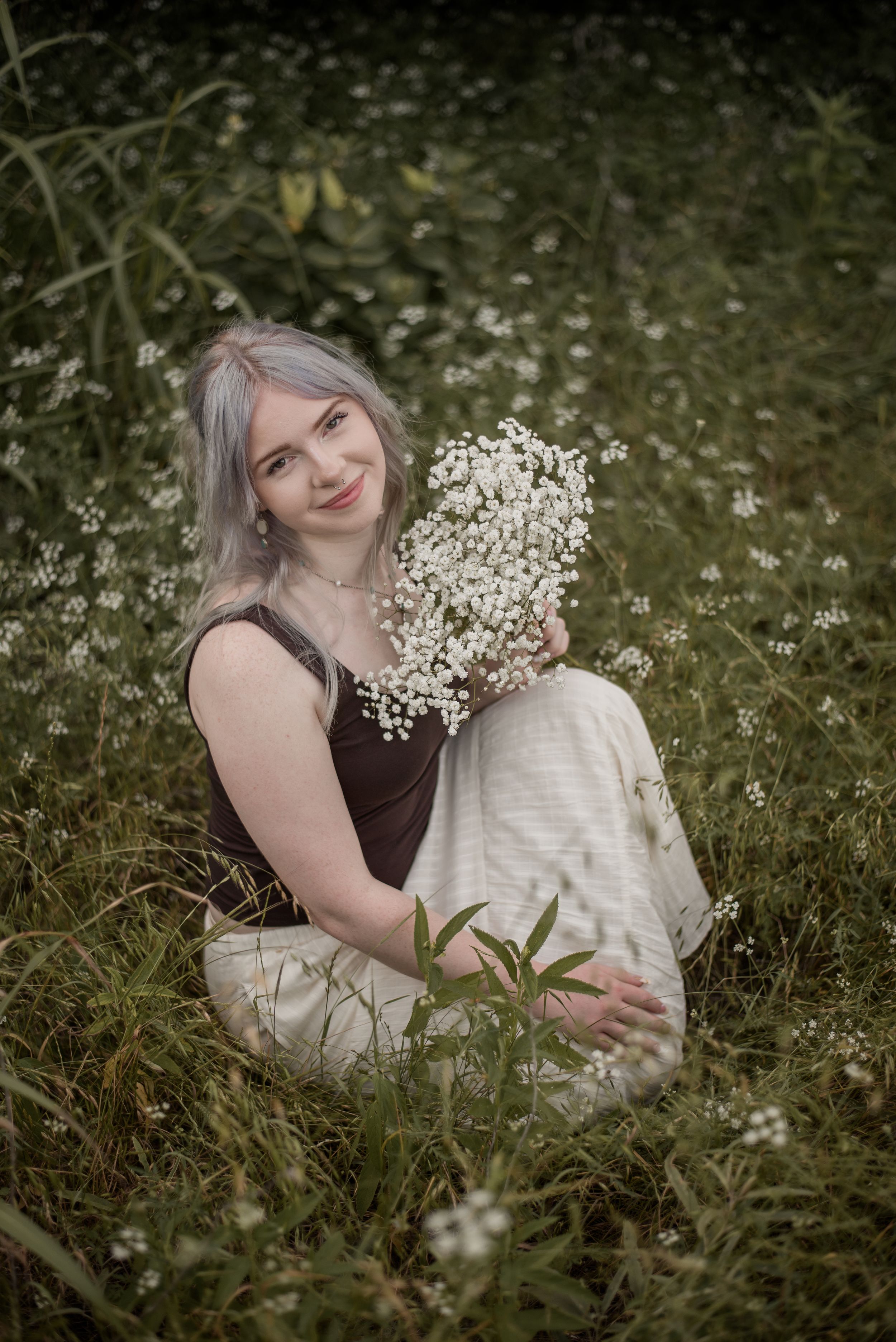 Young woman with silver hair sitting in a wildflower meadow, smiling while holding white baby's breath blooms.