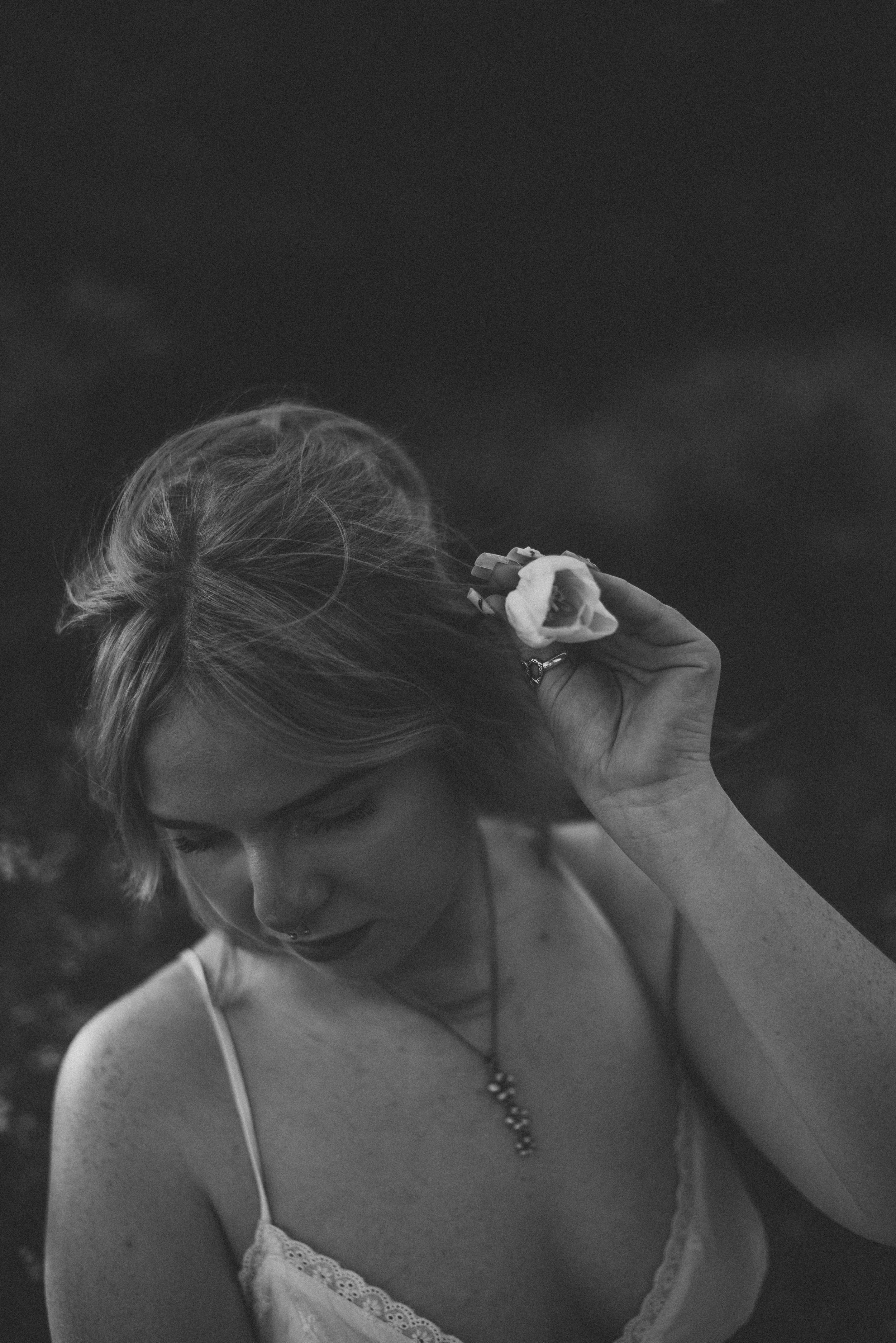 Black and white portrait of a woman holding a flower in her hair while looking down outdoors.