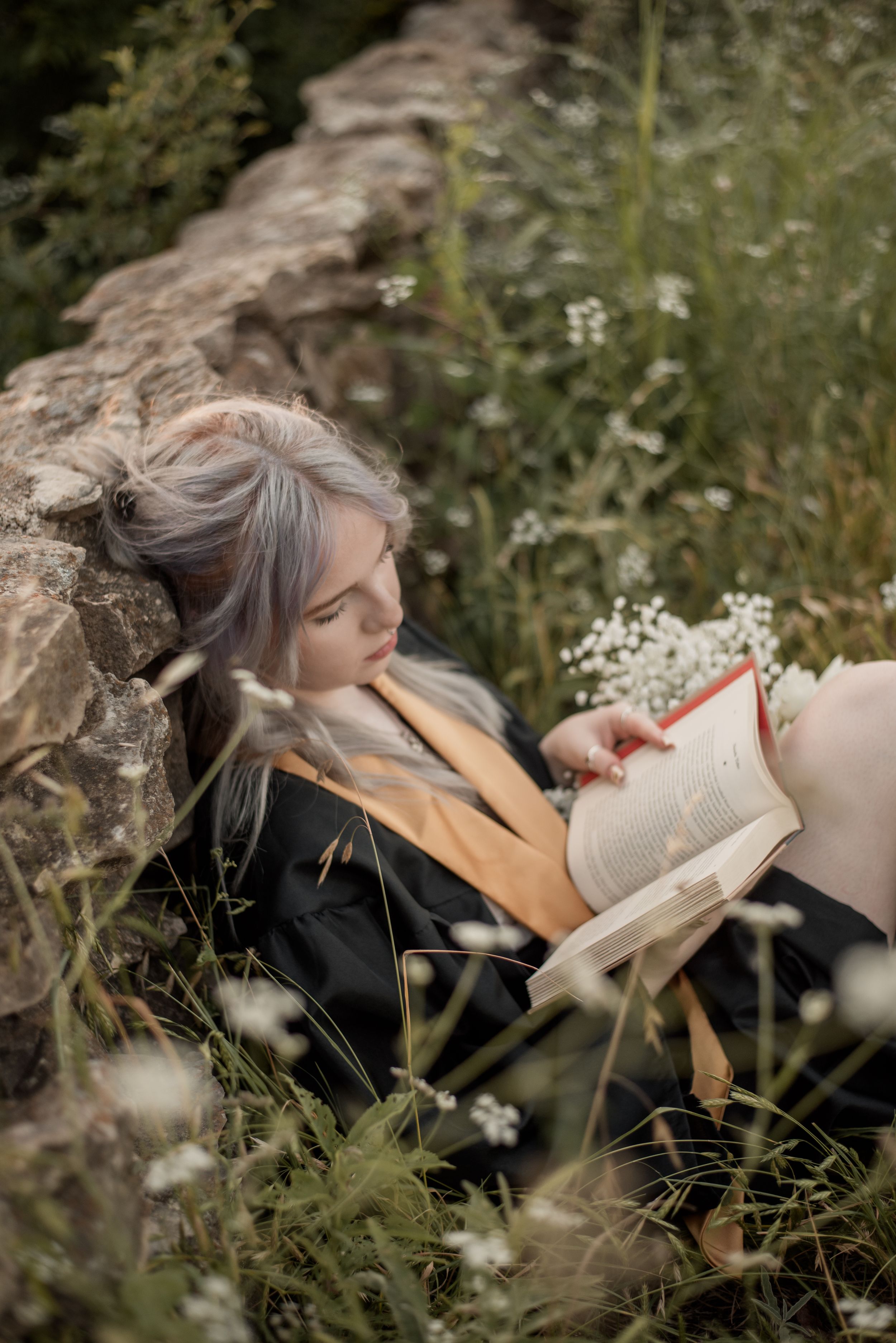 A young woman with silver hair reads a book while relaxing among wildflowers against a stone wall.