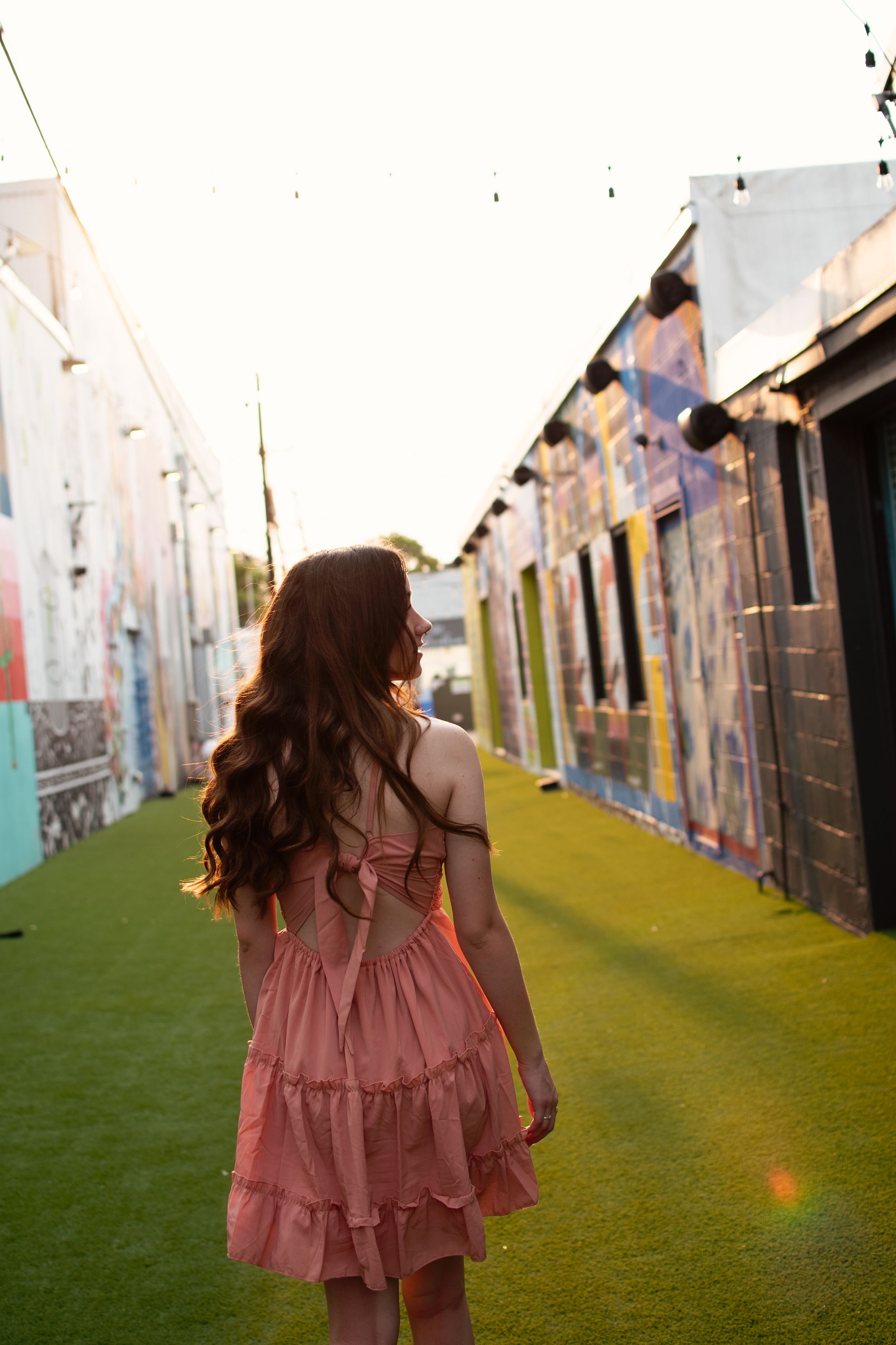 A person in a pink ruffled dress walks down an alley with colorful murals and green turf. Dfw Senior Session