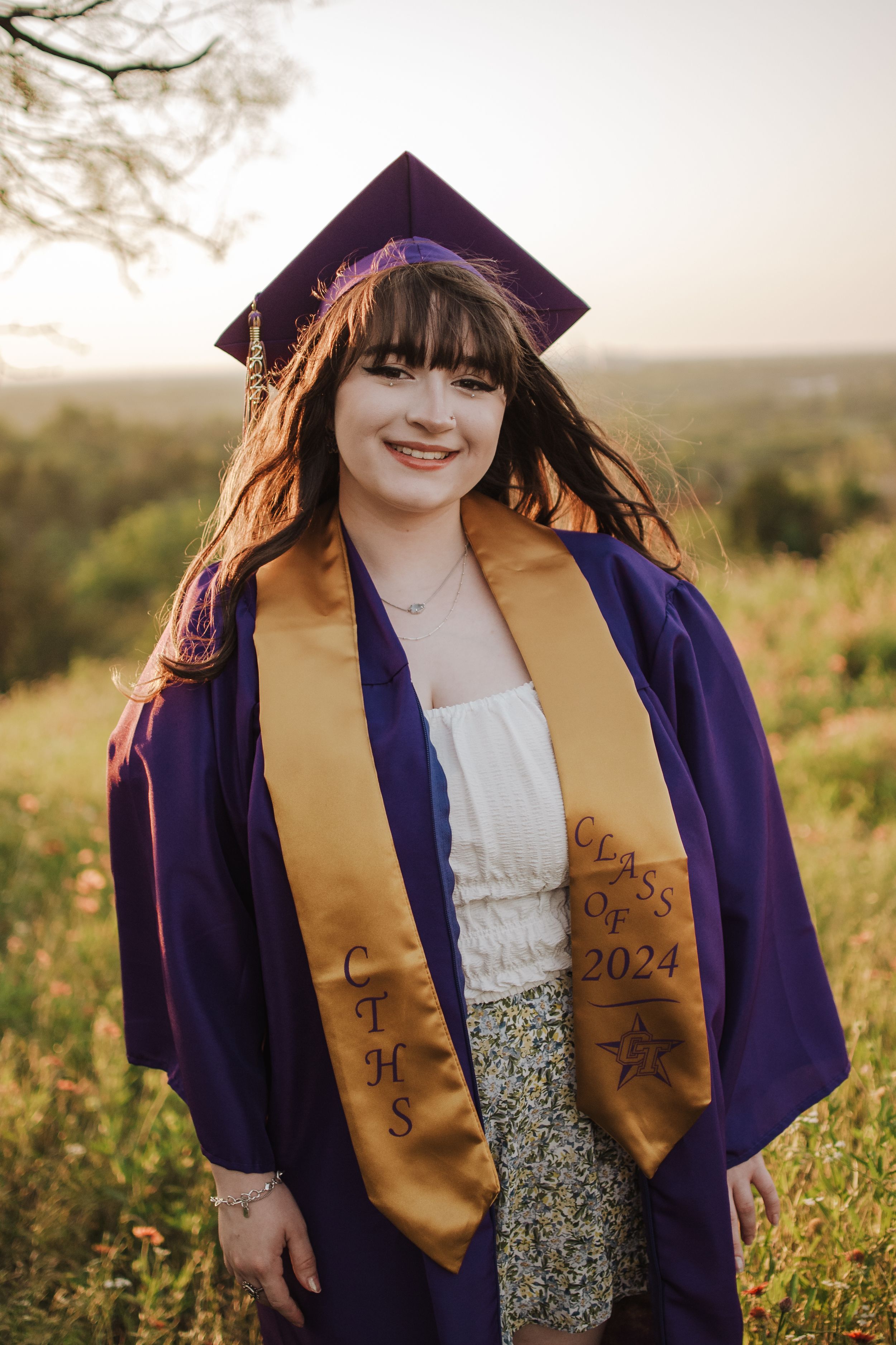 A graduation portrait featuring purple cap and gold stole in a sunset meadow. Tandy Hills