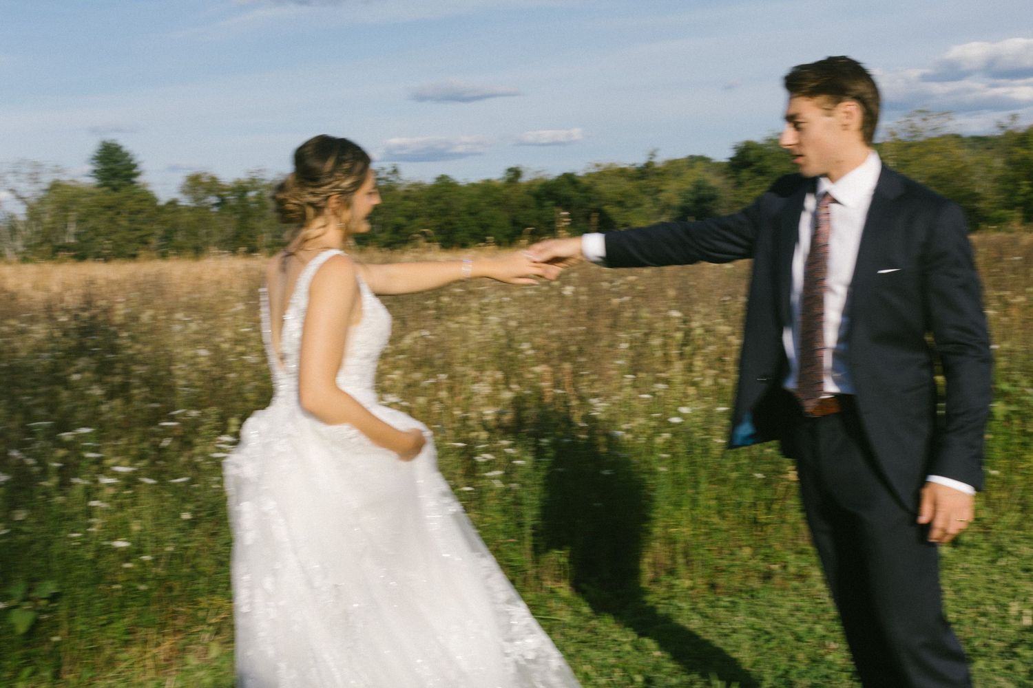 Newlyweds walking hand in hand through a sunny meadow with blue skies overhead.