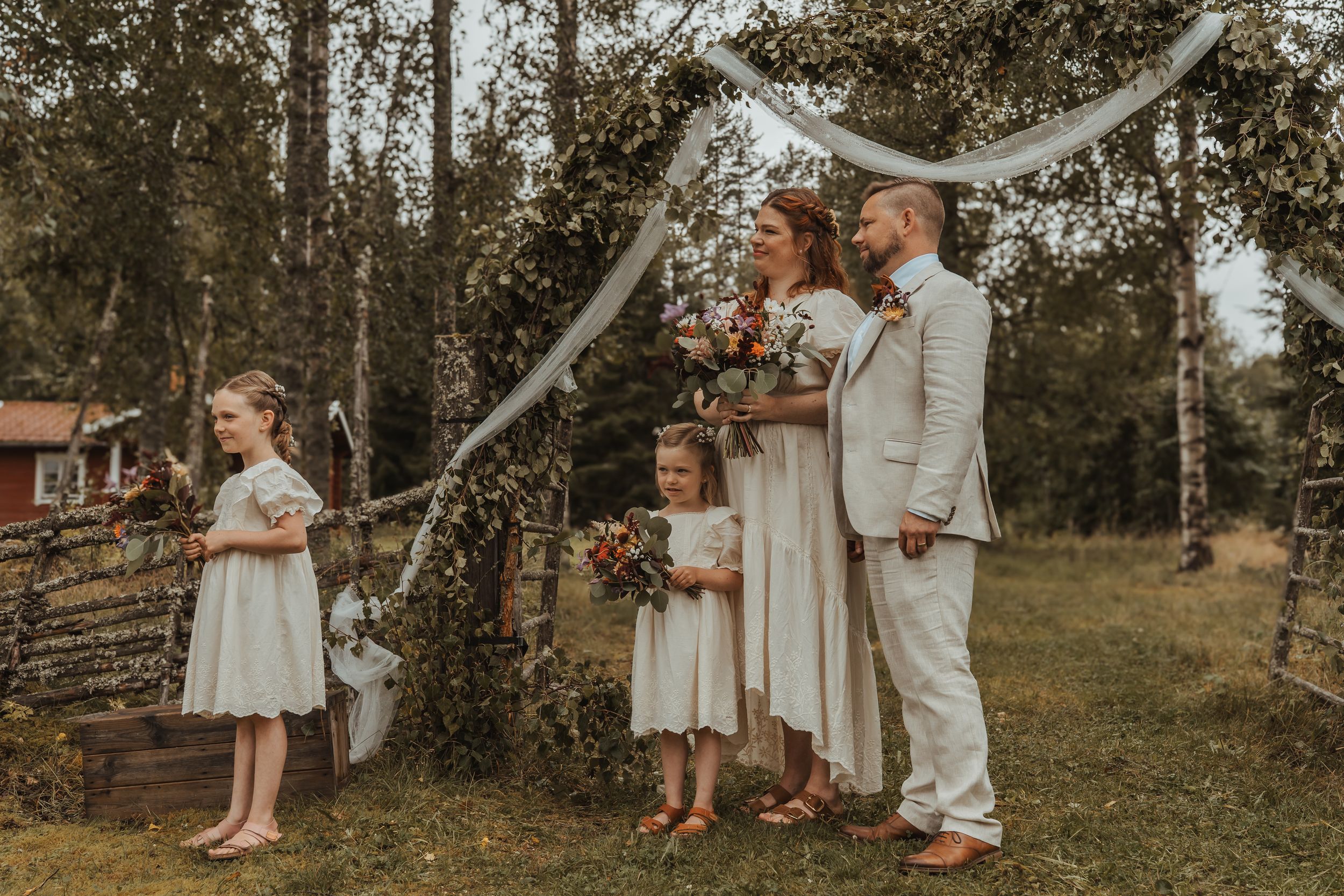 Rustic outdoor wedding scene with flowing white streamers and wedding party in light colored attire against wooded backdrop.