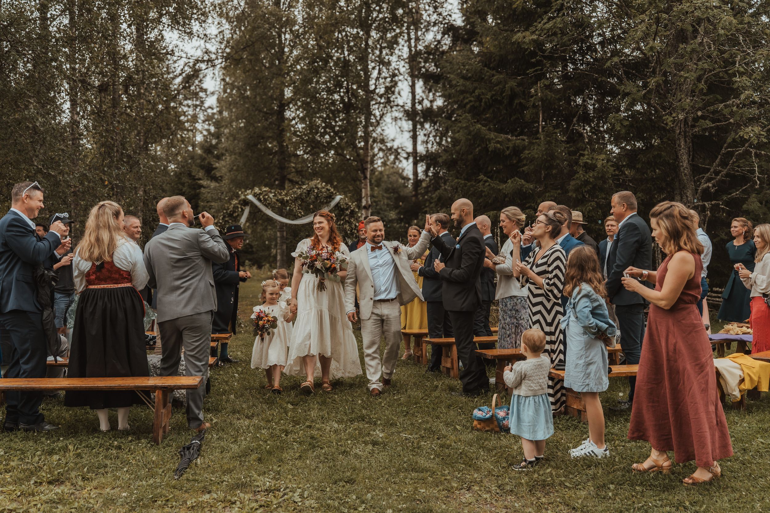A rustic outdoor wedding celebration with guests gathered around wooden benches in a forest clearing.