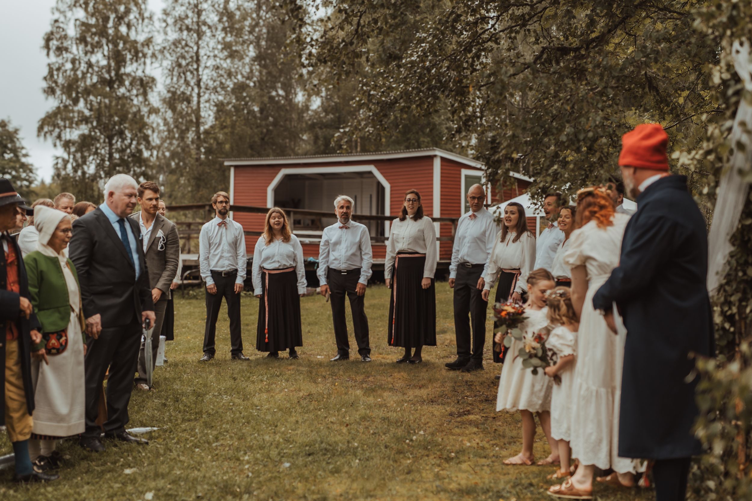 Rustic outdoor wedding ceremony group gathered on green lawn in front of a red barn on autumn day.