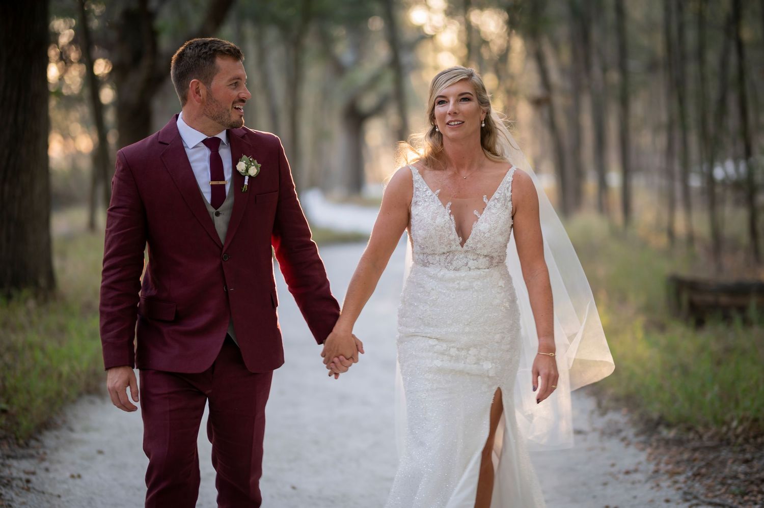 A couple in wedding attire walks through a wooded area during golden hour sunset light.