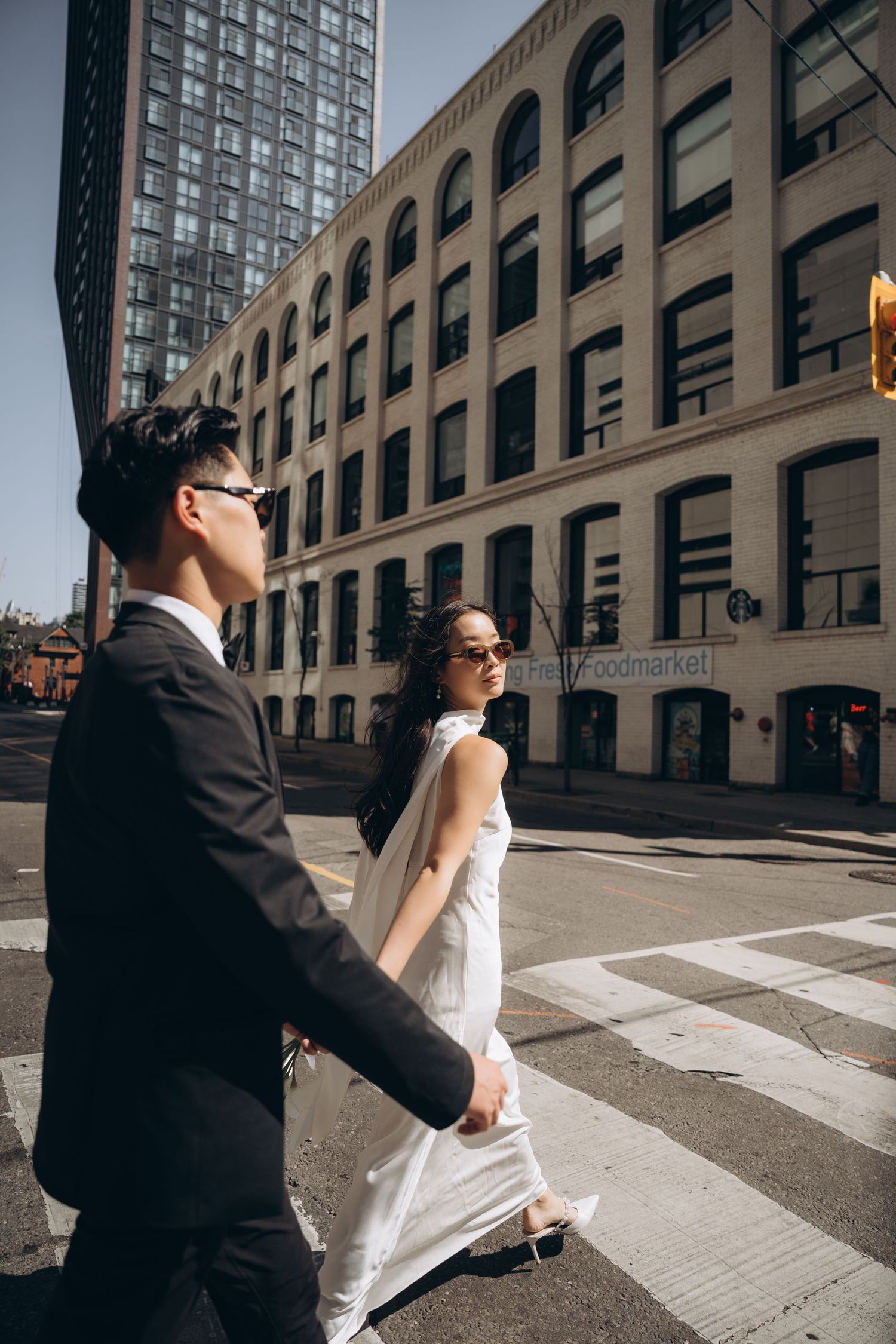 A couple dressed formally crosses an urban crosswalk with classic architecture and tall buildings in downtown setting.