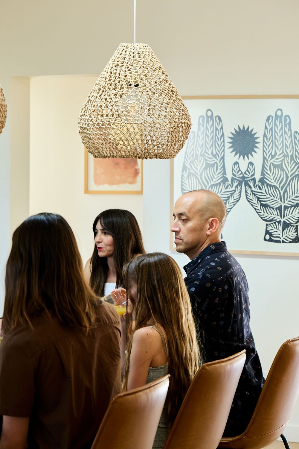 Interior view of dining area with a woven pendant lamp and people gathered beneath artwork featuring hands and a sun motif.