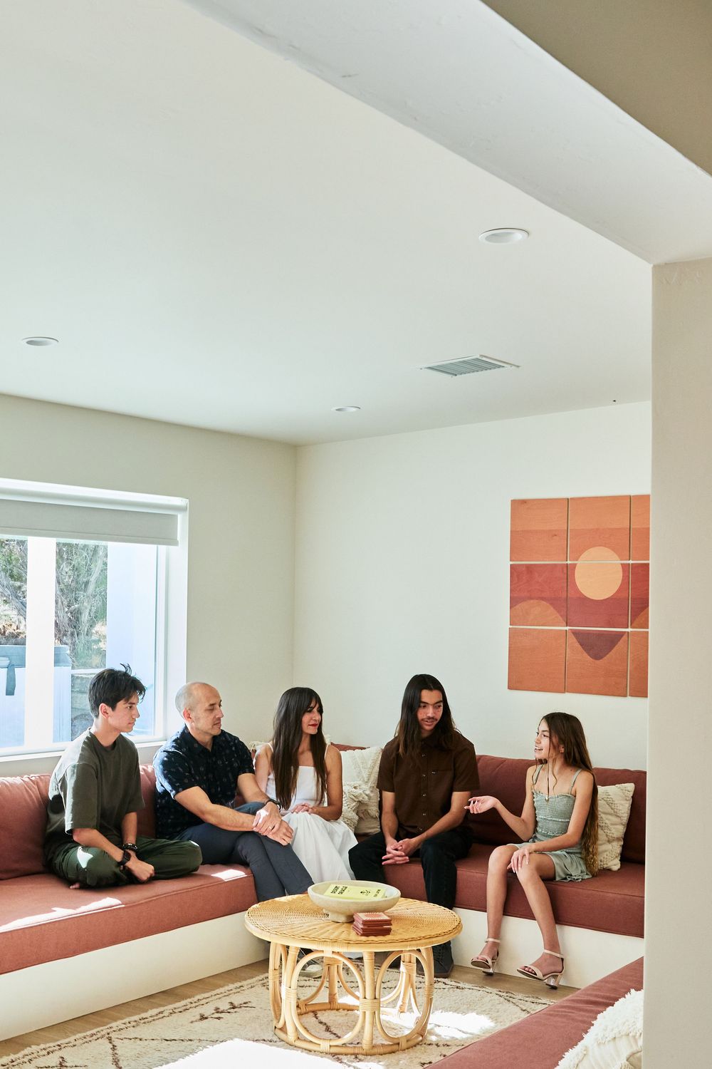 A group of people sitting together on a built-in bench in a modern living room with retro orange wall art and rattan table.