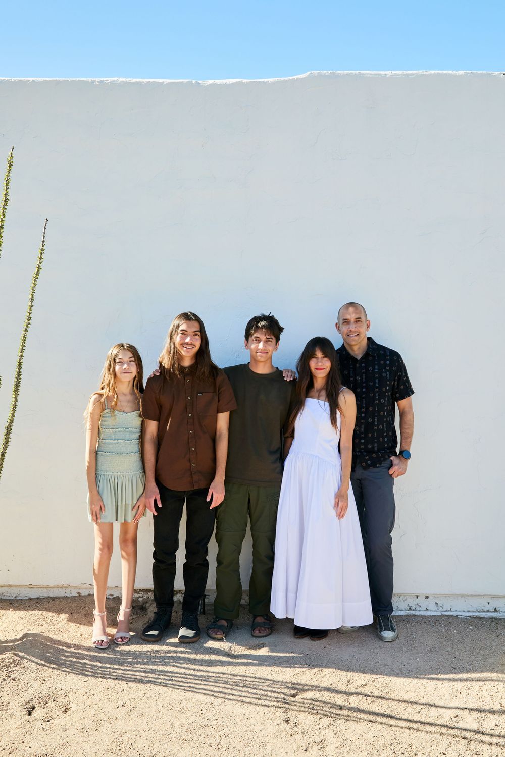 A group of people standing against a white wall outdoors on a sunny day.