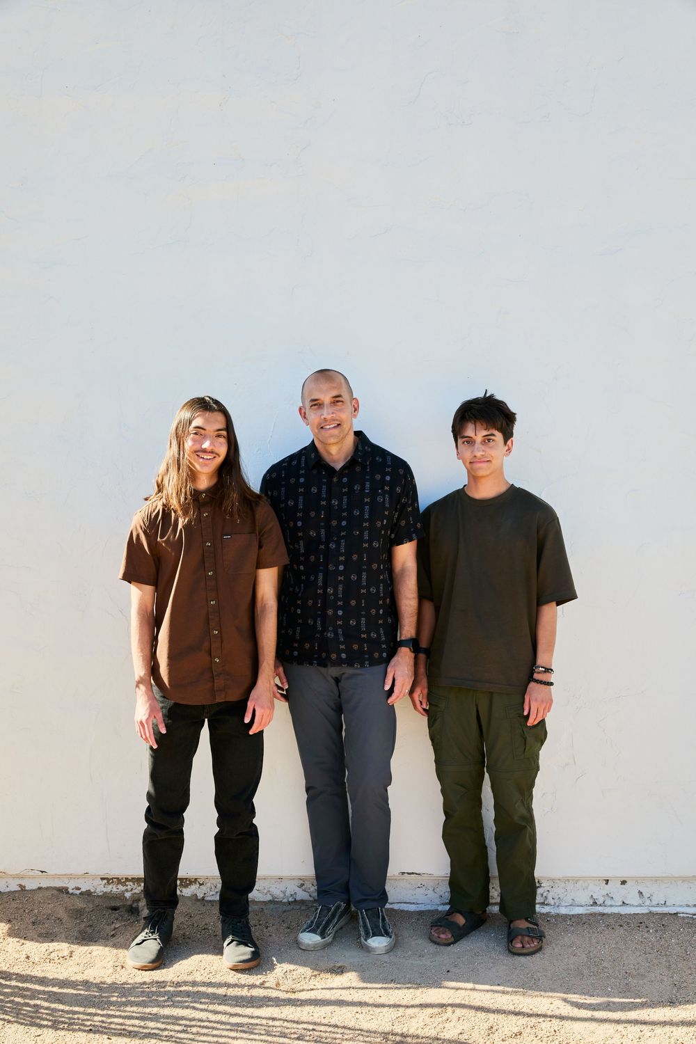 Three people stand against a light wall wearing casual clothing in brown and dark tones.