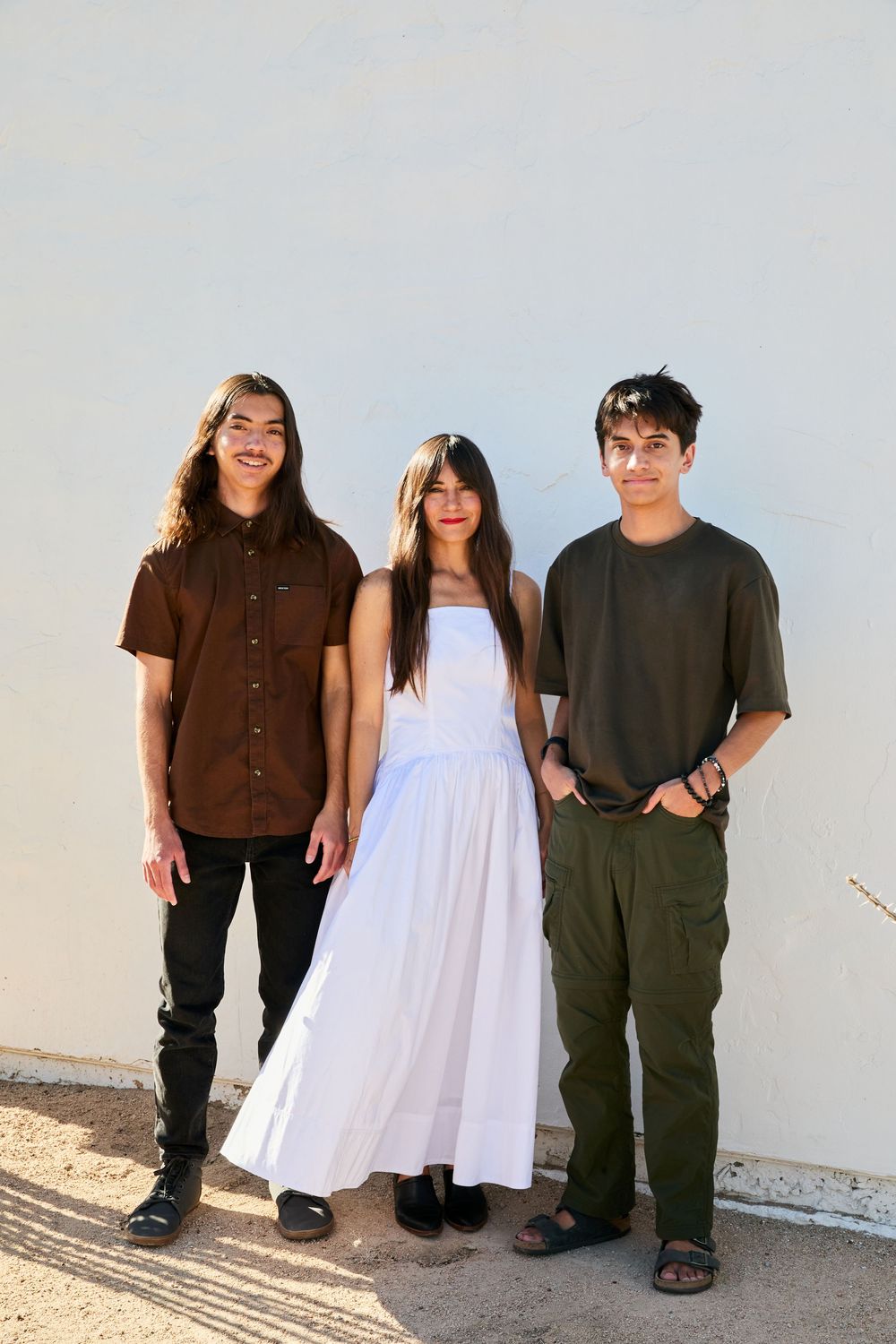 Three people on white wall background wearing casual outfits including white dress and dark clothing.