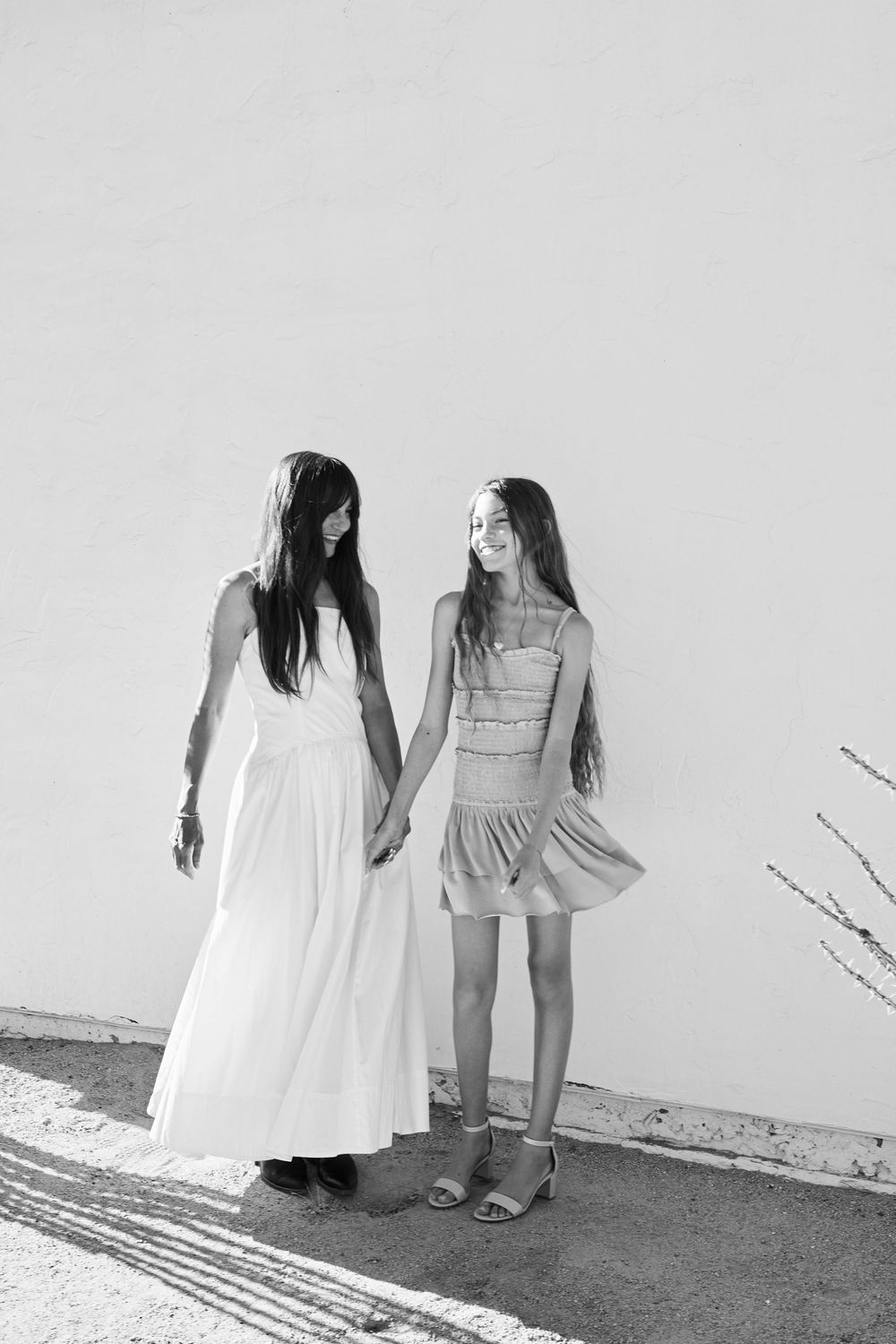 Black and white photo of two friends in dresses standing against a white wall holding hands.