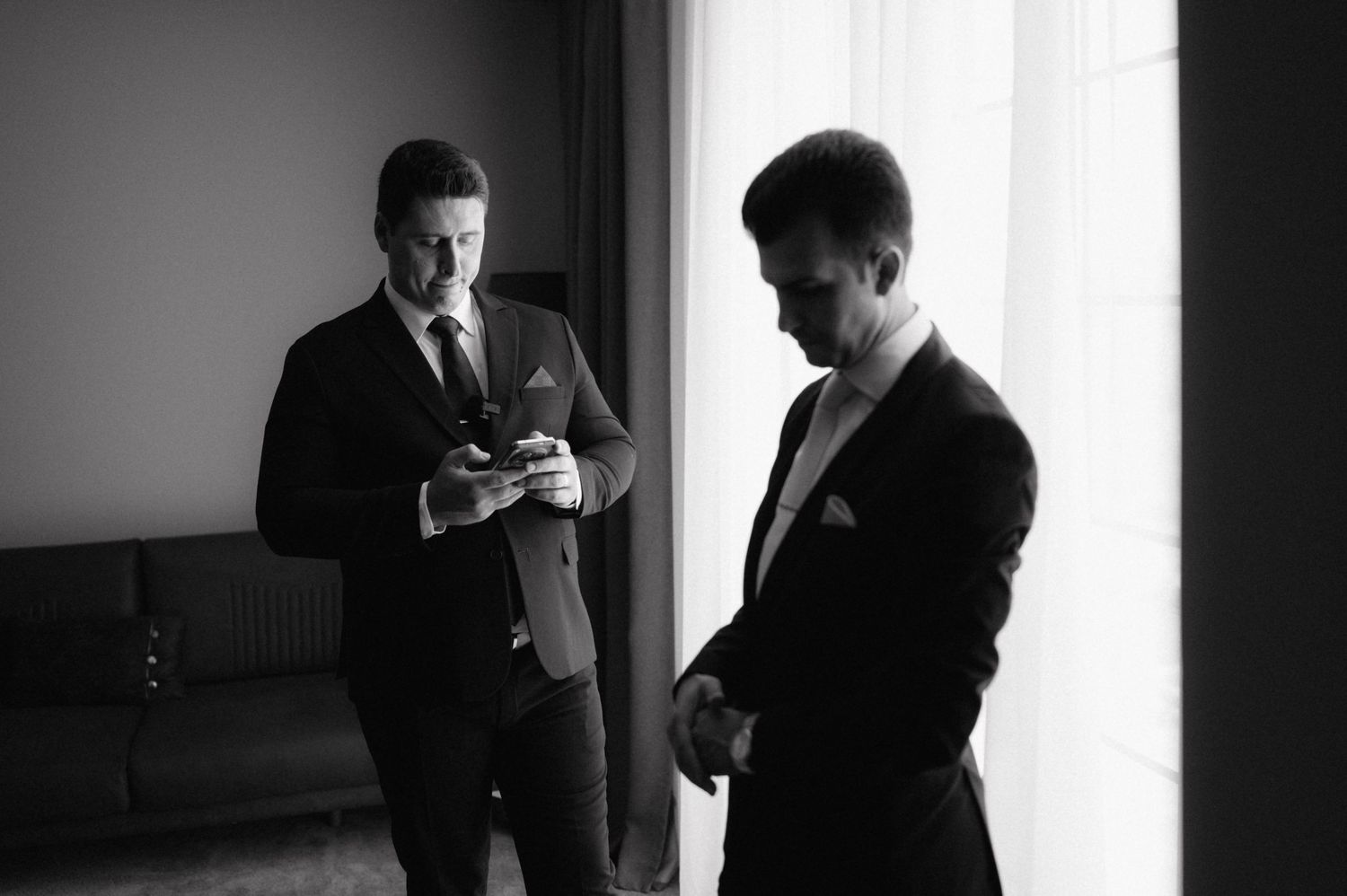Two men in dark suits standing indoors checking their phones in black and white photography.
