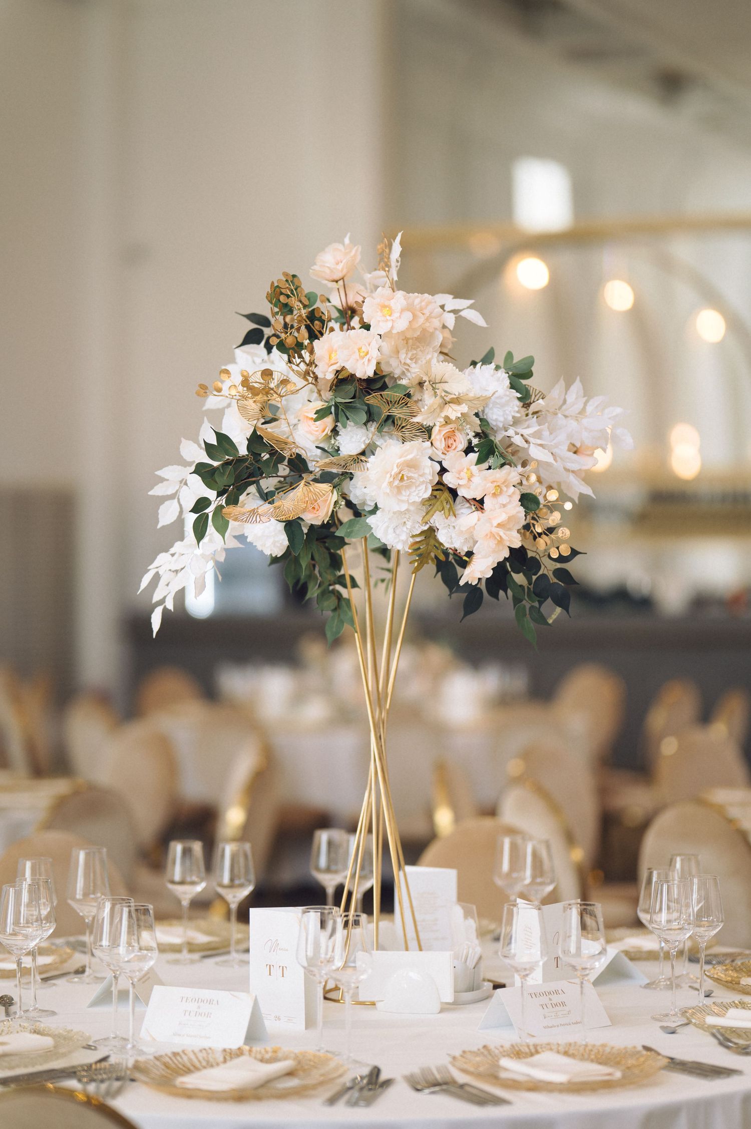 Elegant wedding reception tables with white floral centerpieces in gold vases and formal place settings arranged in a banquet hall.