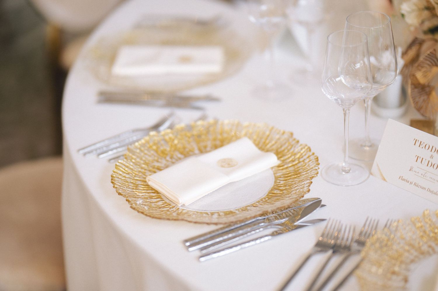 Elegant place setting with gold-rimmed charger plate and white napkin on round wedding reception table.