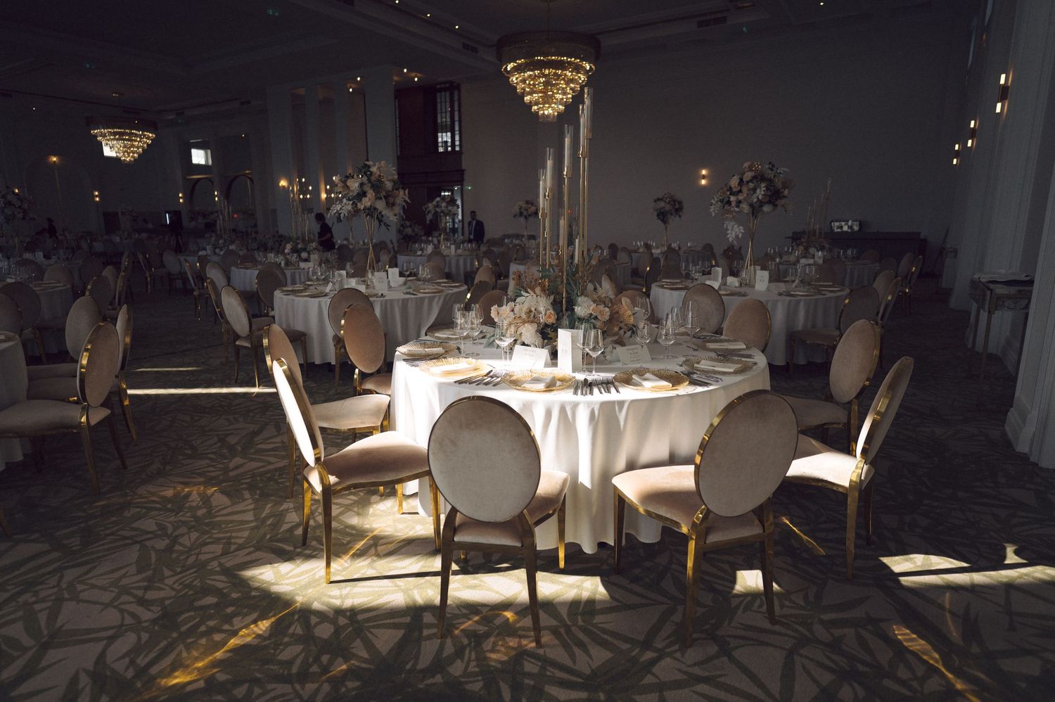 Elegant banquet hall with spotlit round tables, ornate chairs, and crystal chandeliers in a dimly lit atmosphere.
