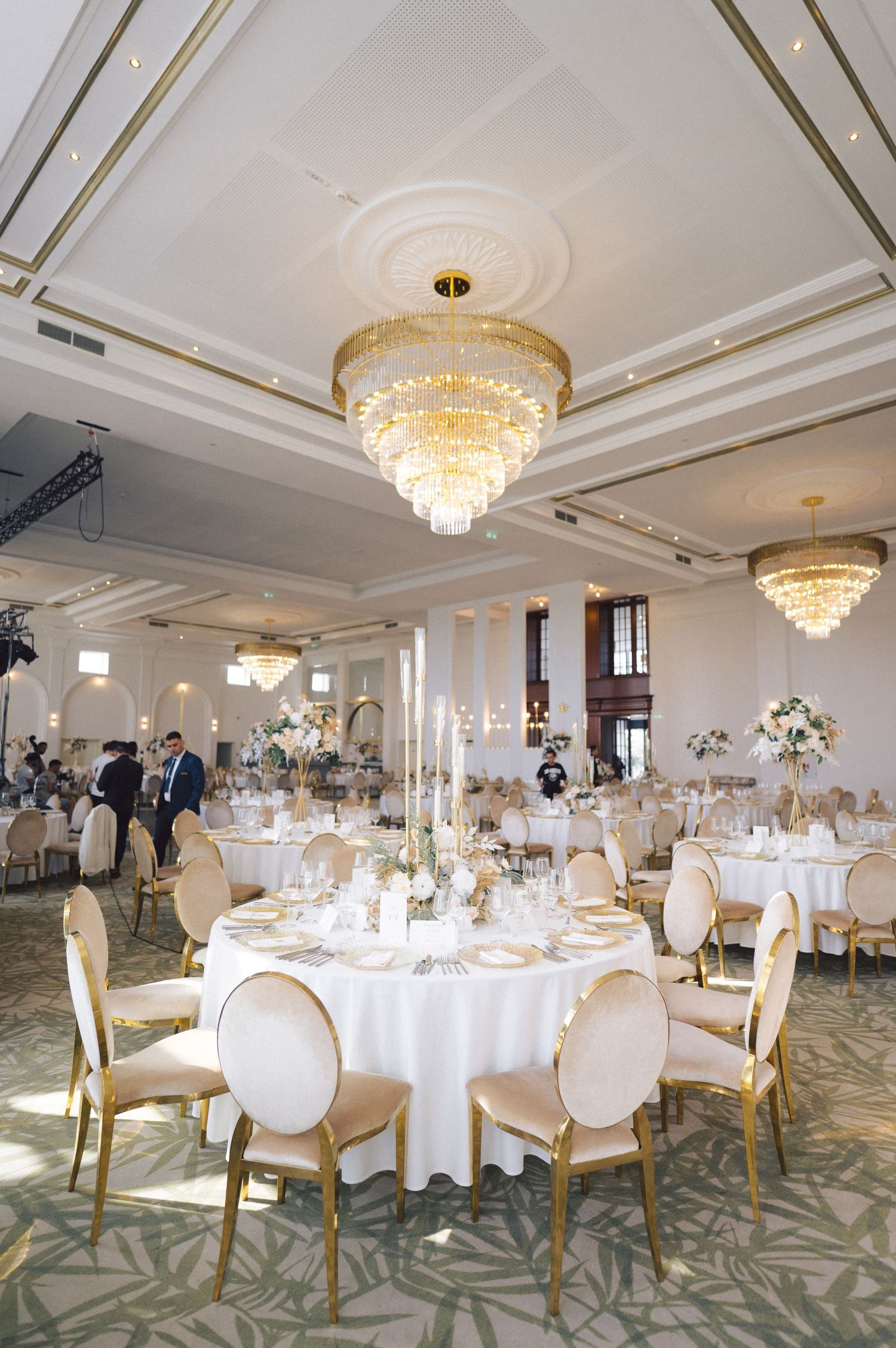Elegant ballroom setup with round tables, white linens, golden chairs, and crystal chandeliers for a luxurious wedding reception.