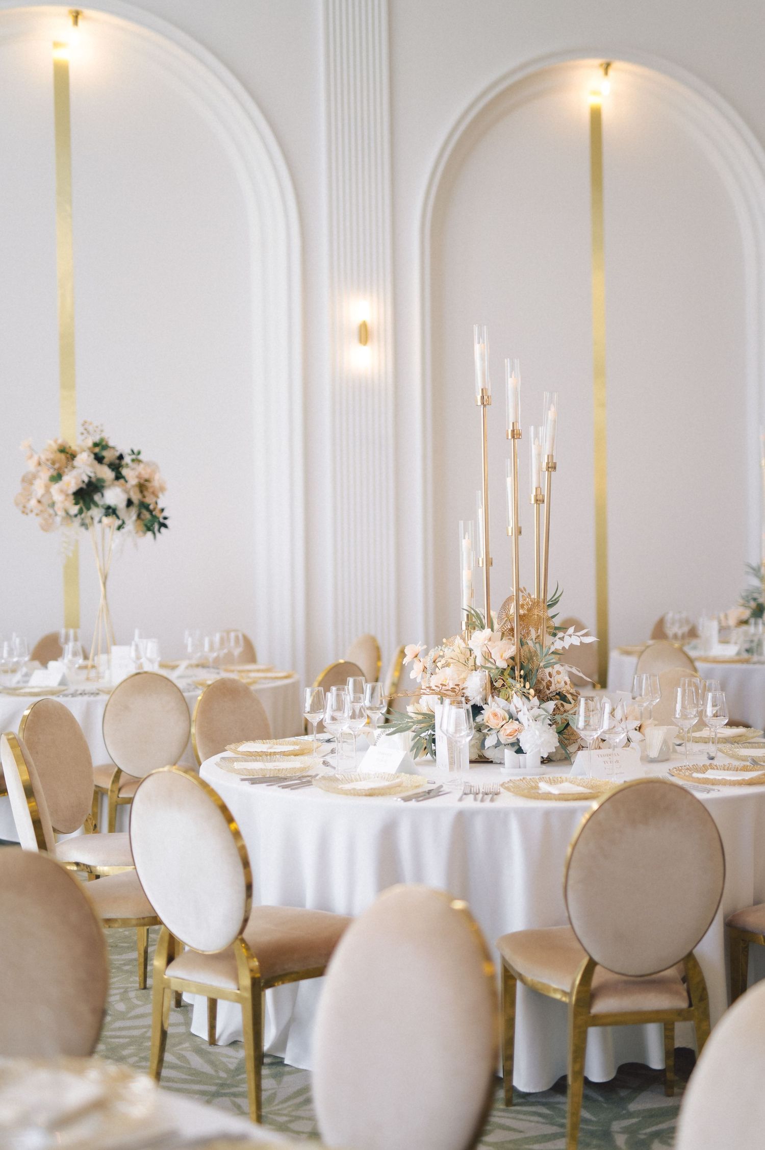 Elegant white wedding reception venue with round tables, cream chairs, tall candles and floral centerpieces under arched walls.