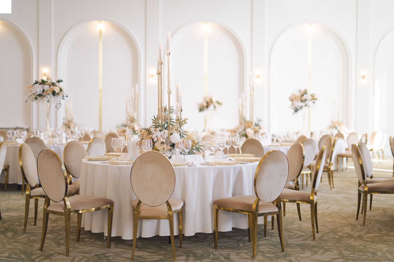 Elegant wedding reception venue with white round tables, French-style chairs, and floral arrangements under arched windows.