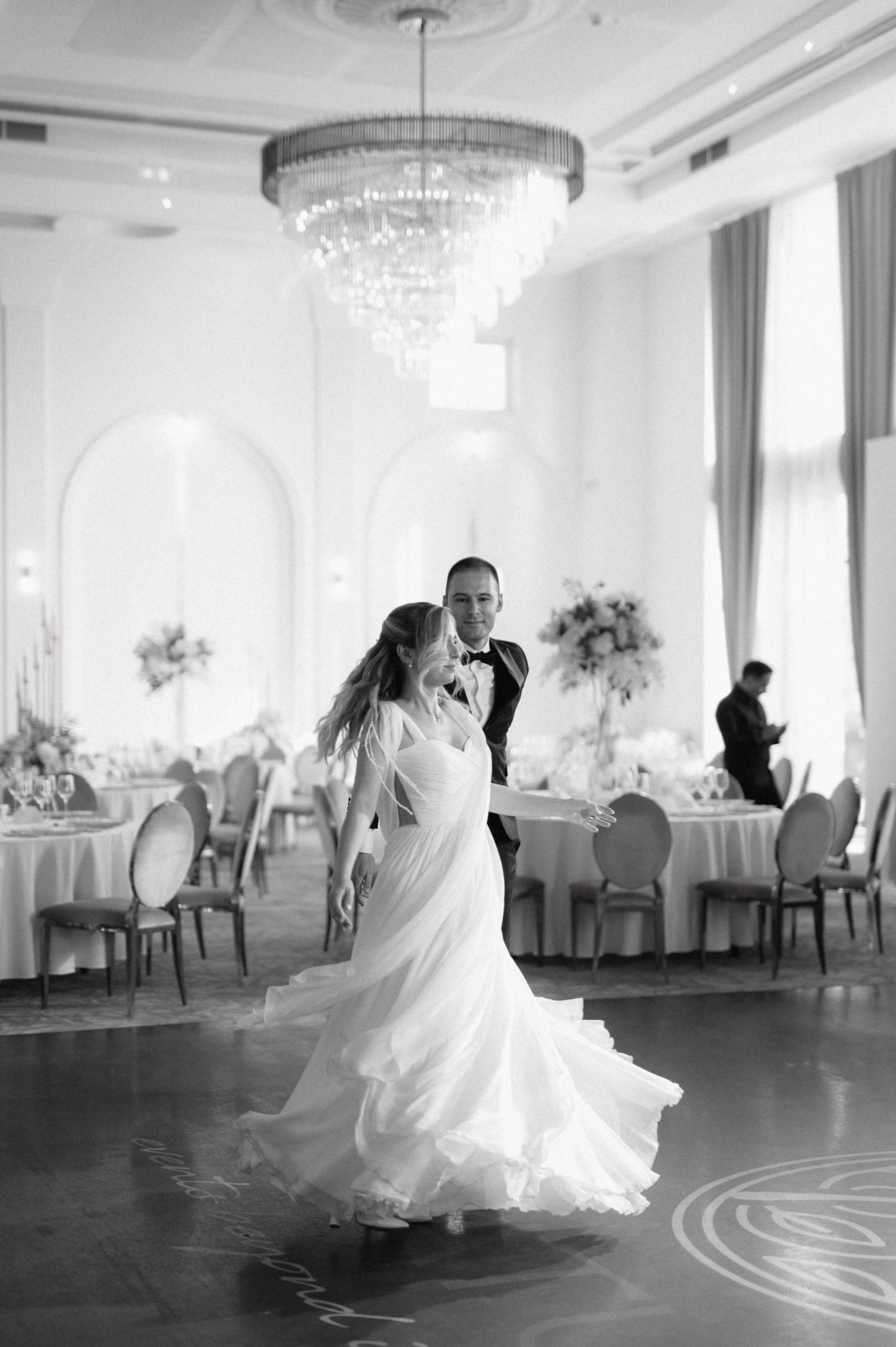 A couple dancing in an elegant white ballroom with crystal chandelier and round dining tables.