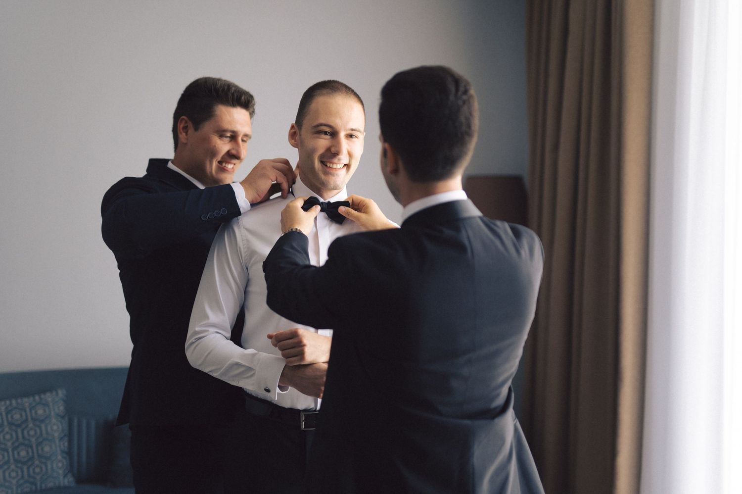 Groomsmen help adjust formal attire while getting ready in a hotel room with natural window lighting.