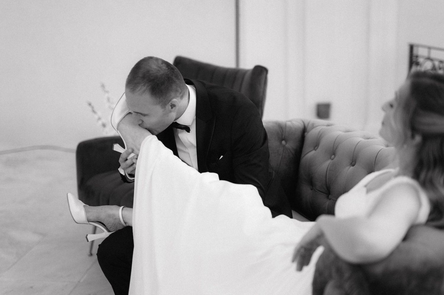 A black and white intimate wedding photograph shows a quiet emotional moment on a tufted couch.