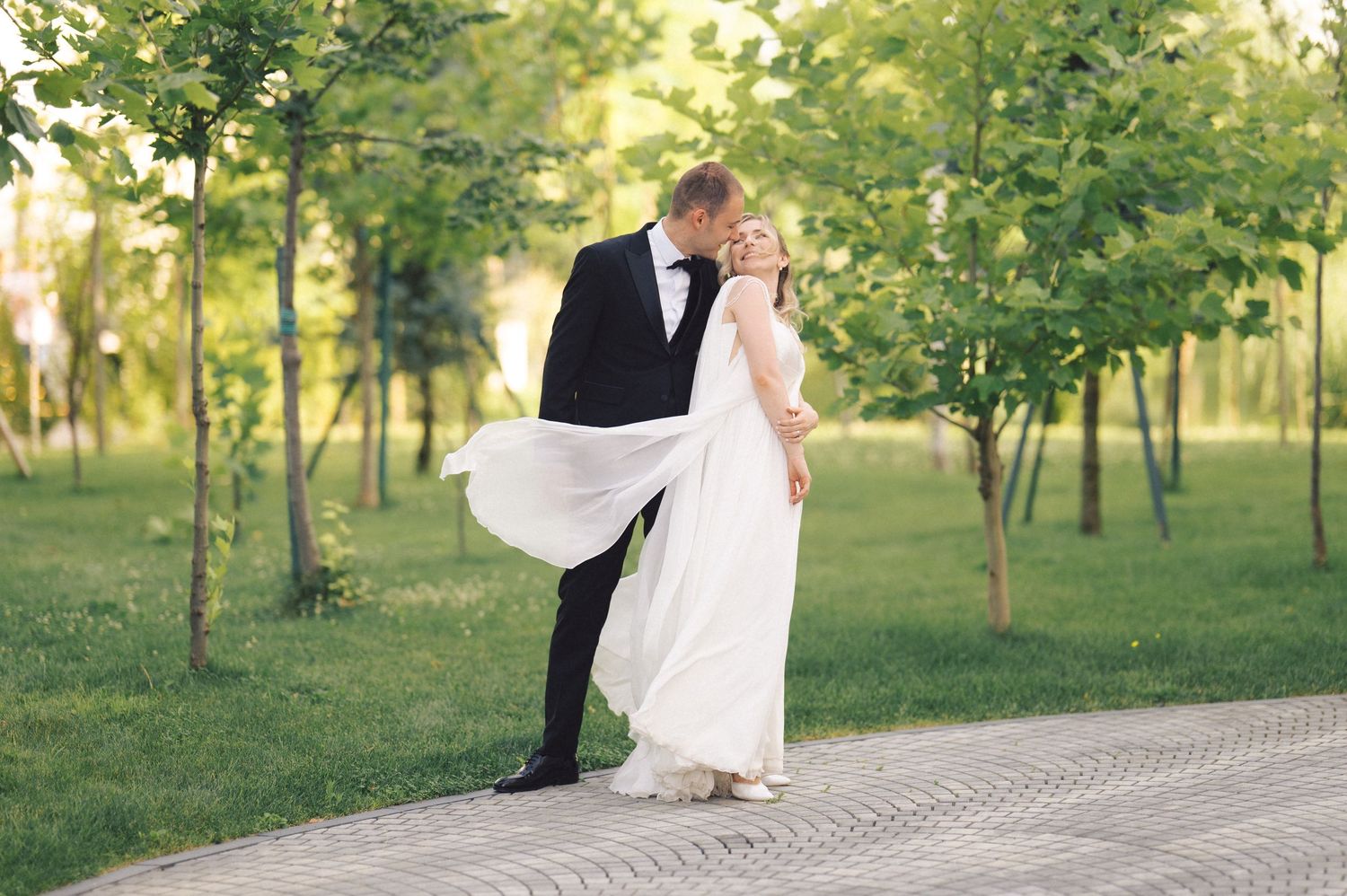 A couple in formal wedding attire walk together on a path surrounded by green trees in soft sunlight.