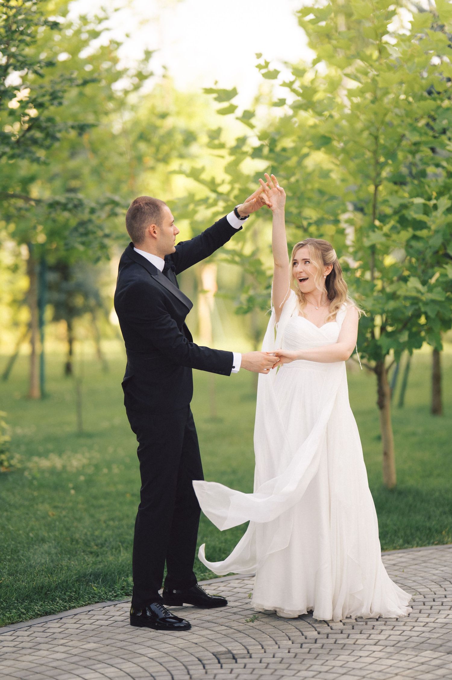 A couple dances joyfully together on a stone path surrounded by green trees in a romantic outdoor setting.