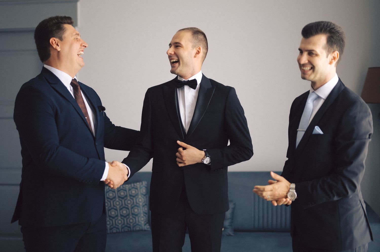 Groomsmen in dark suits smile and share a lighthearted moment while getting ready at a formal wedding.
