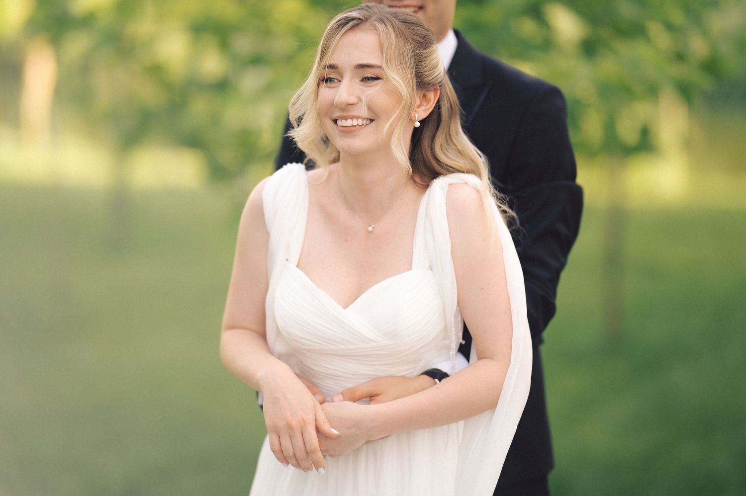 A bride in a white dress smiles joyfully during an outdoor wedding ceremony in a sunlit garden setting.