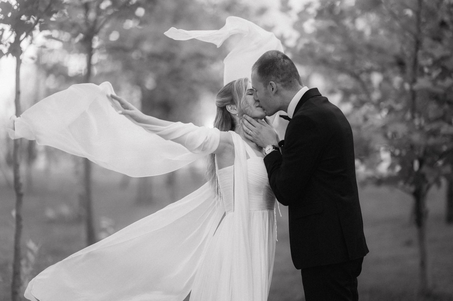 A romantic black and white wedding photo capturing a couple kissing as the bride's veil flows dramatically in the wind.