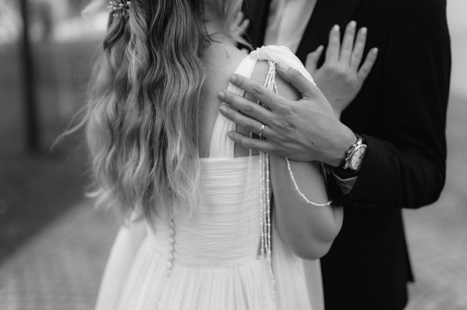 A black and white photograph shows a person in a dark suit holding the hands of someone in a white dress.
