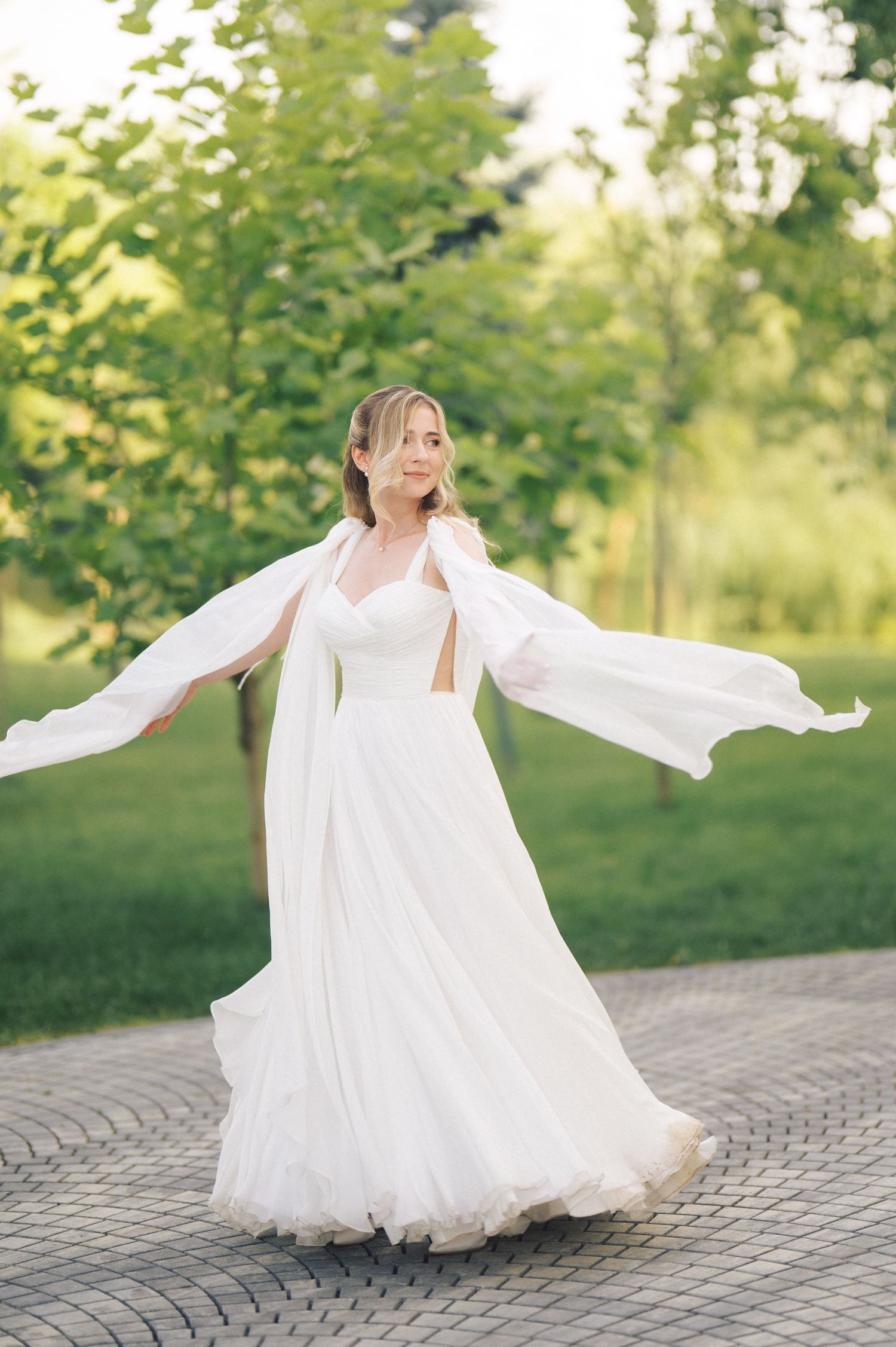 A bride in a flowing white dress with long bell sleeves twirls on a brick paver path surrounded by lush greenery.
