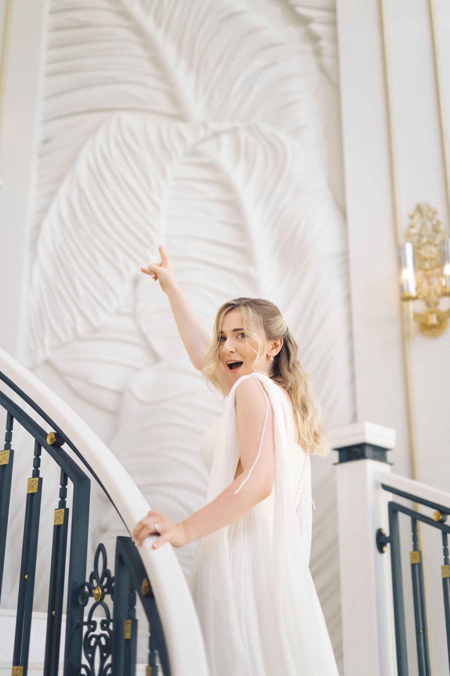 Figure in a flowing white dress poses elegantly on an ornate staircase with palm leaf wall decor.