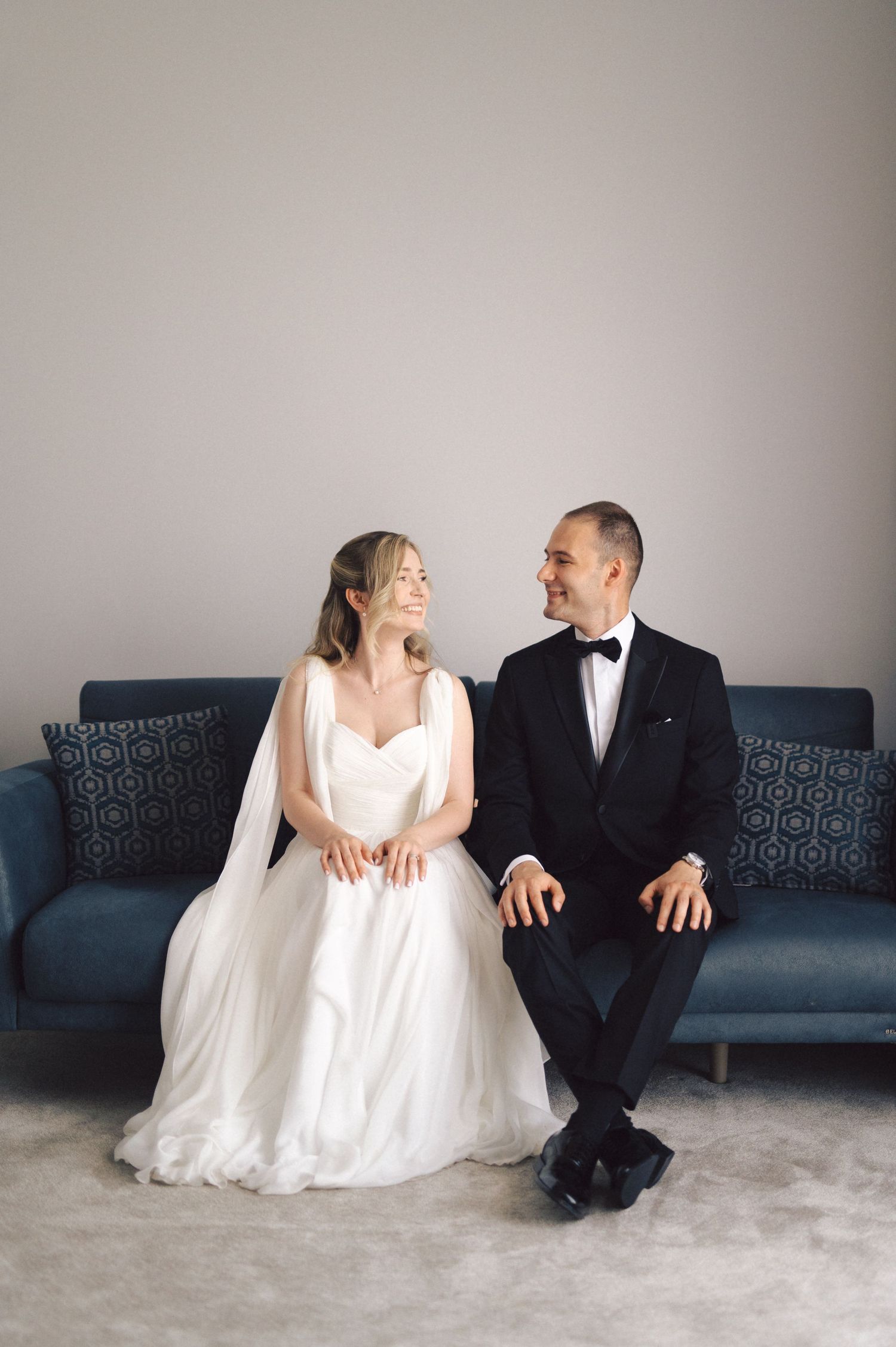 A bride and groom share a joyful moment while sitting together on a navy blue couch against a neutral wall.