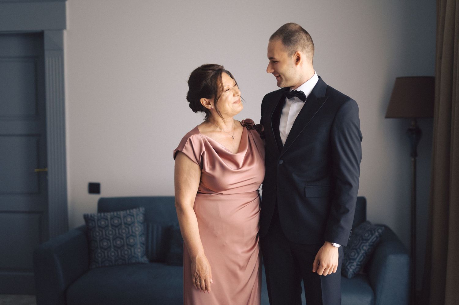 A tender moment between two people in formal attire, with one wearing a dusky pink gown and the other in a black tuxedo.