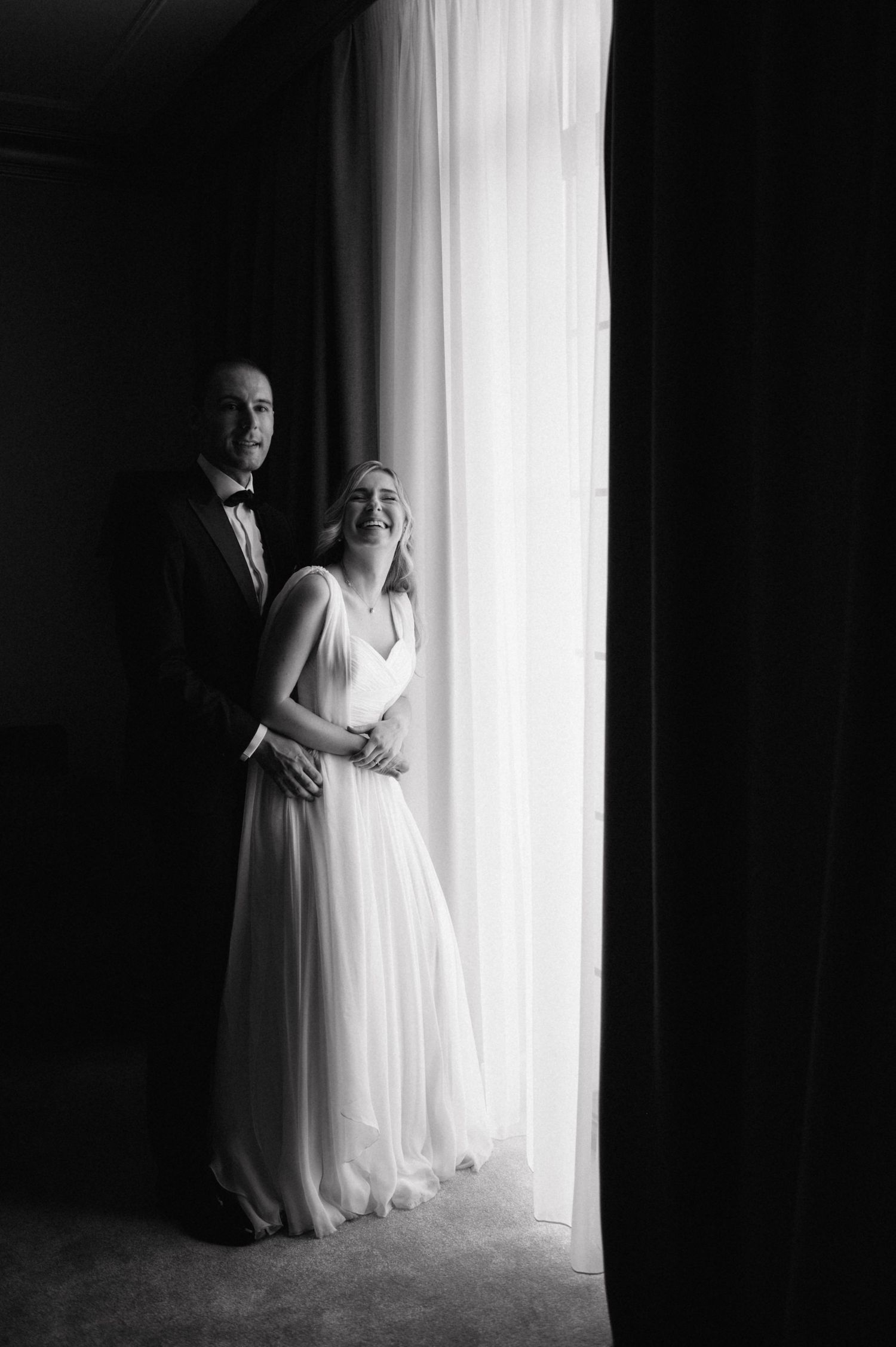 A black and white artistic portrait of a couple by a window in formal wedding attire.