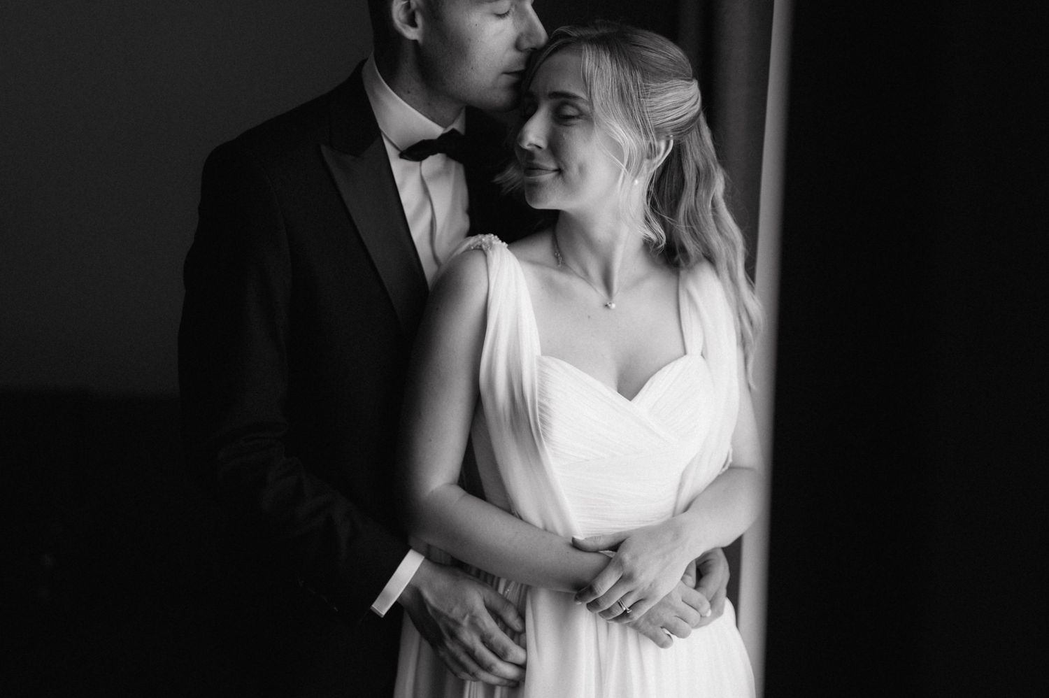 Dramatic black and white wedding portrait captures intimate embrace near window.
