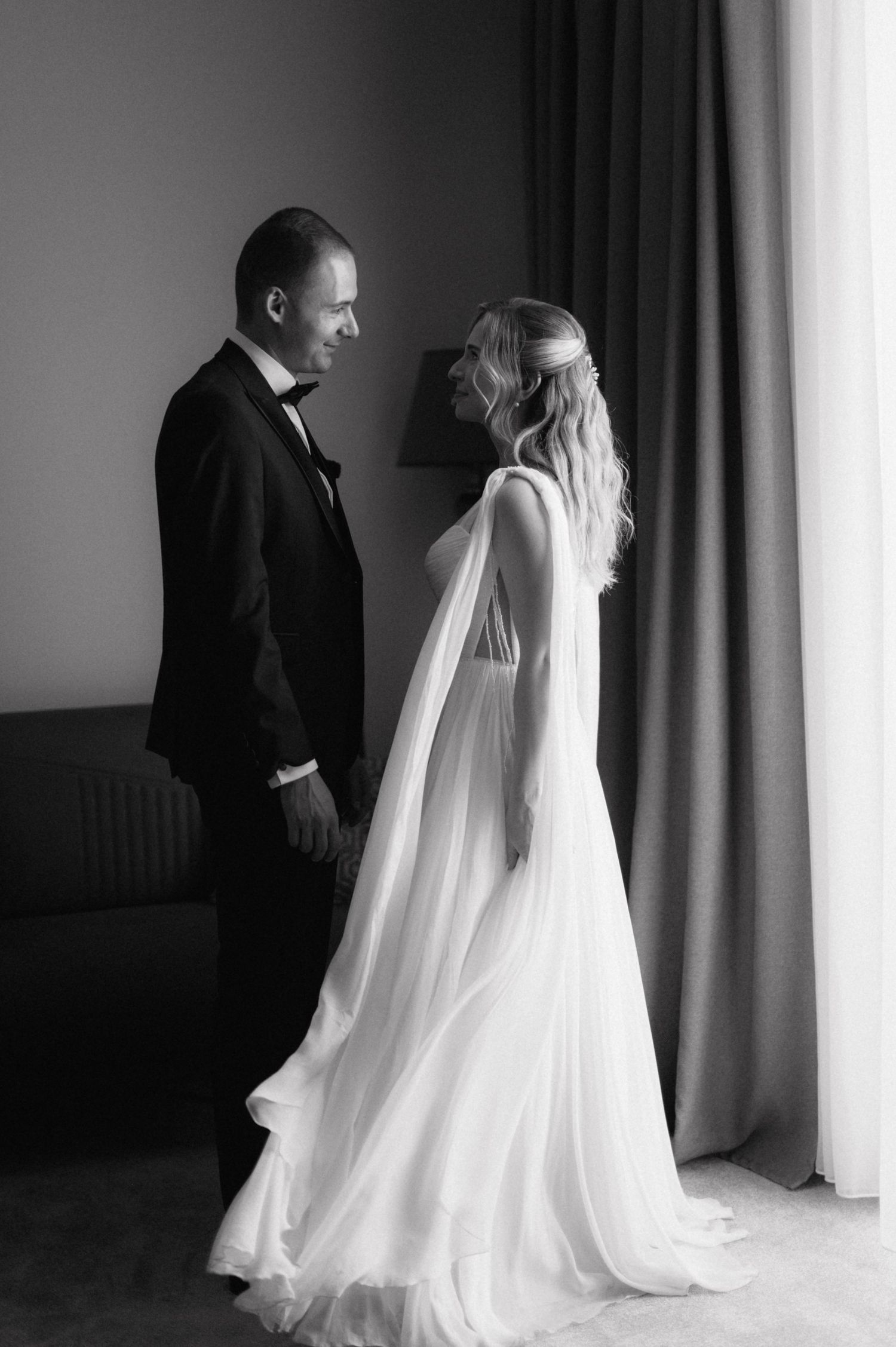 An elegant black and white wedding portrait shows a couple in formal attire standing by curtained windows.