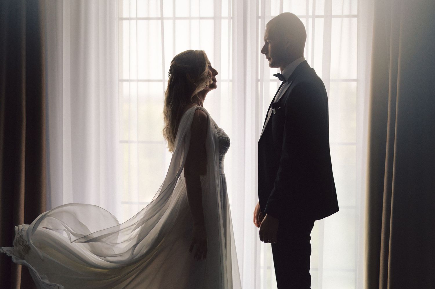 A silhouetted couple in wedding attire standing together by a bright window with flowing veil.