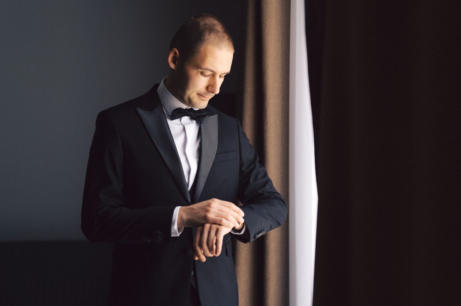 A person wearing a black tuxedo and bow tie checks their watch while standing by a window in moody lighting.
