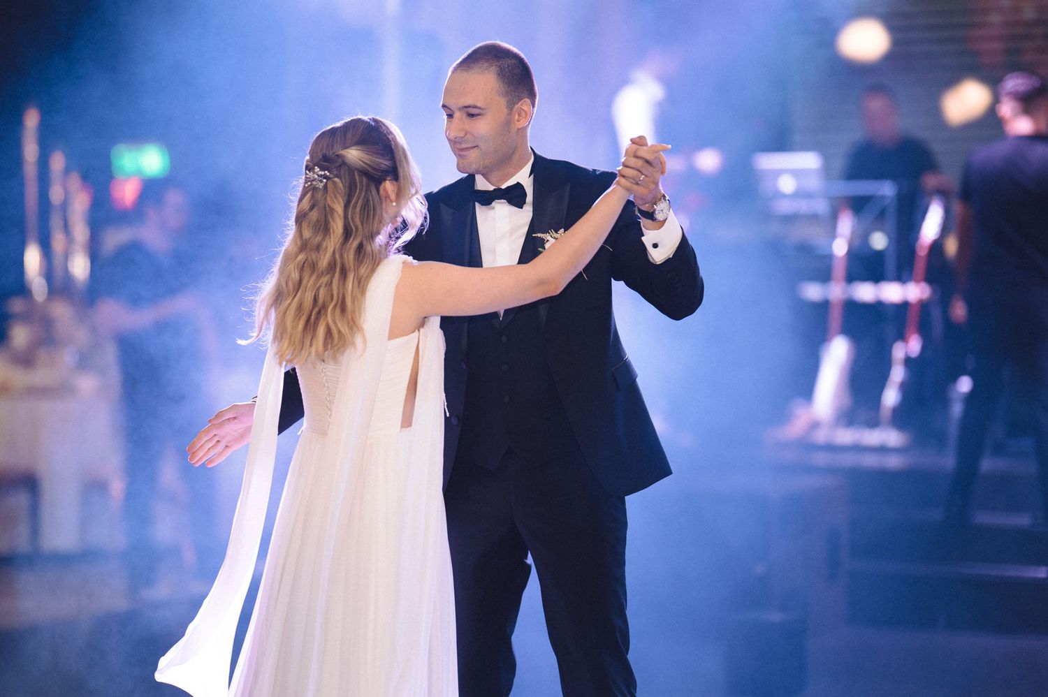 A couple in formal attire dances their first dance at a wedding reception amid soft lighting and fog effects.