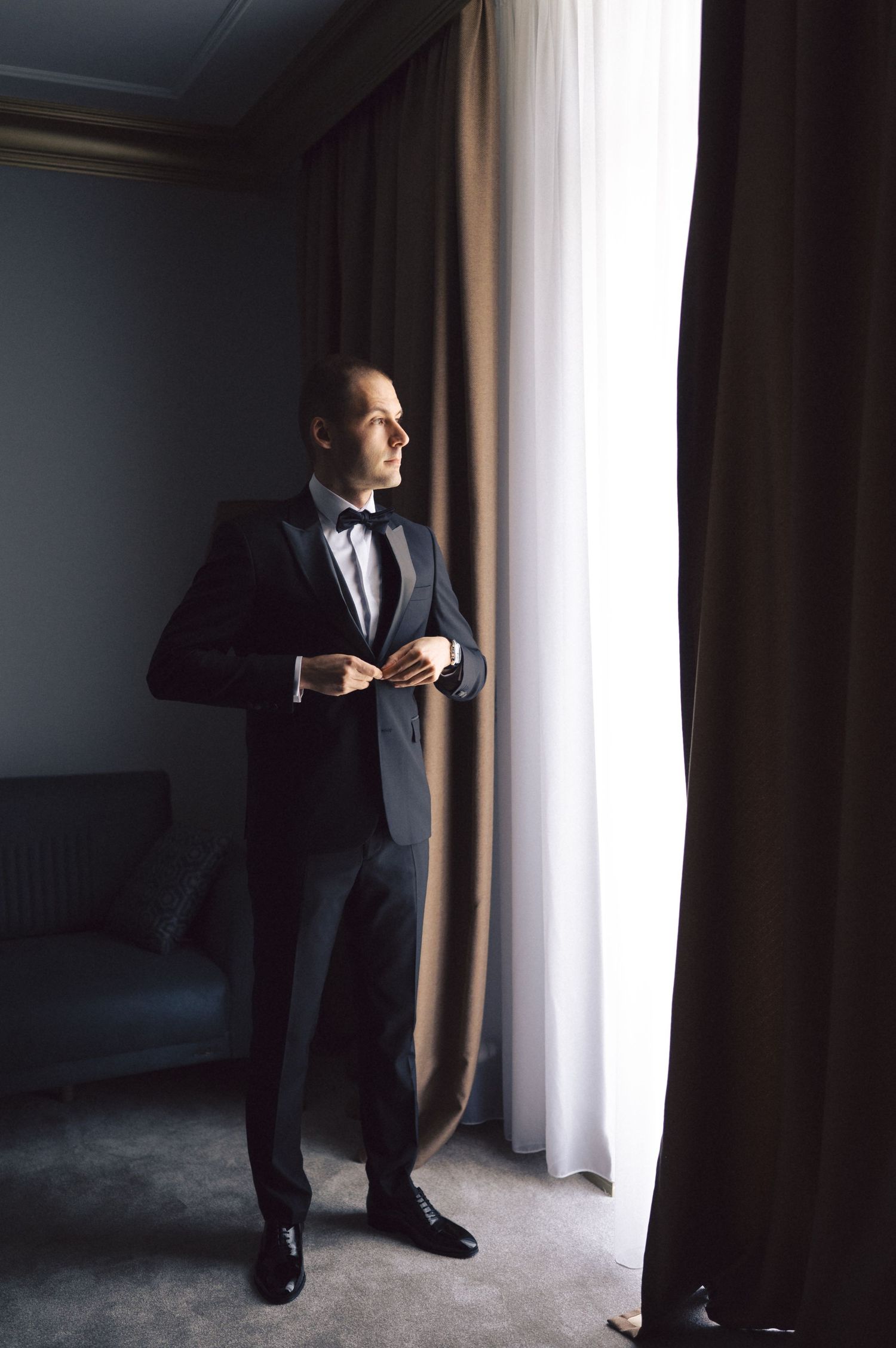 A silhouetted figure in a dark suit stands by a curtained window in a moody, atmospheric interior.